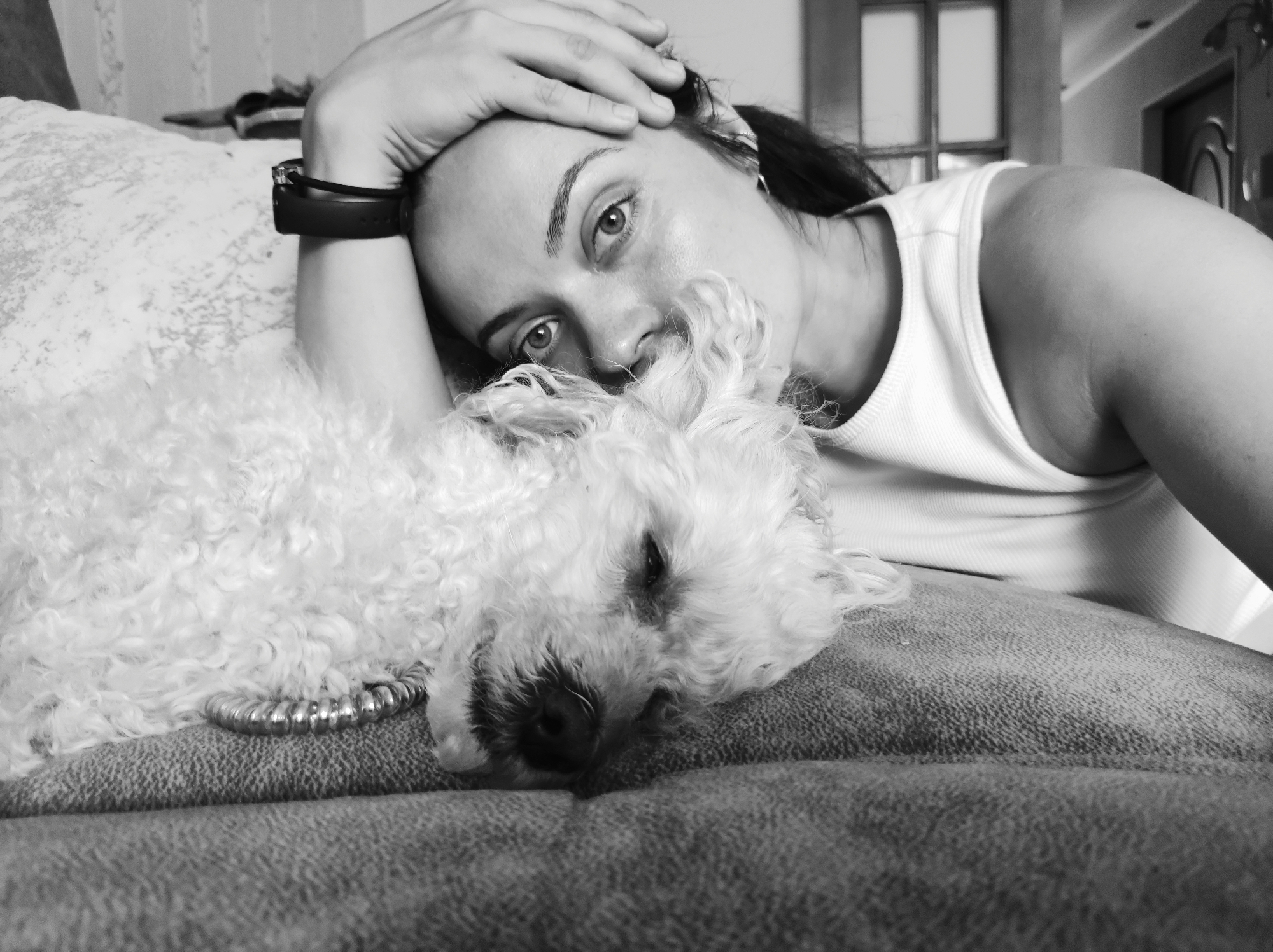 In a black and white, eye-level shot, a woman with dark hair rests her head on her hand, her arm bent, as she looks directly at the camera. Her gaze is intense, with her eyes wide open and her eyebrows slightly raised. She is wearing a white tank top and a black fitness tracker on her wrist. Beside her, a small, white, fluffy dog, possibly a poodle, is asleep with its head resting on the woman's arm and its snout partially obscured by her hair. The dog wears a metallic, segmented collar. They are both positioned on a textured, couch-like surface. The background is out of focus, but hints of indoor elements like a window frame and a doorway are visible. The scene appears to be a relaxed, intimate moment, likely in a home environment. The image does not provide any specific clues to place it in the city of Chaikovsky, Russia.