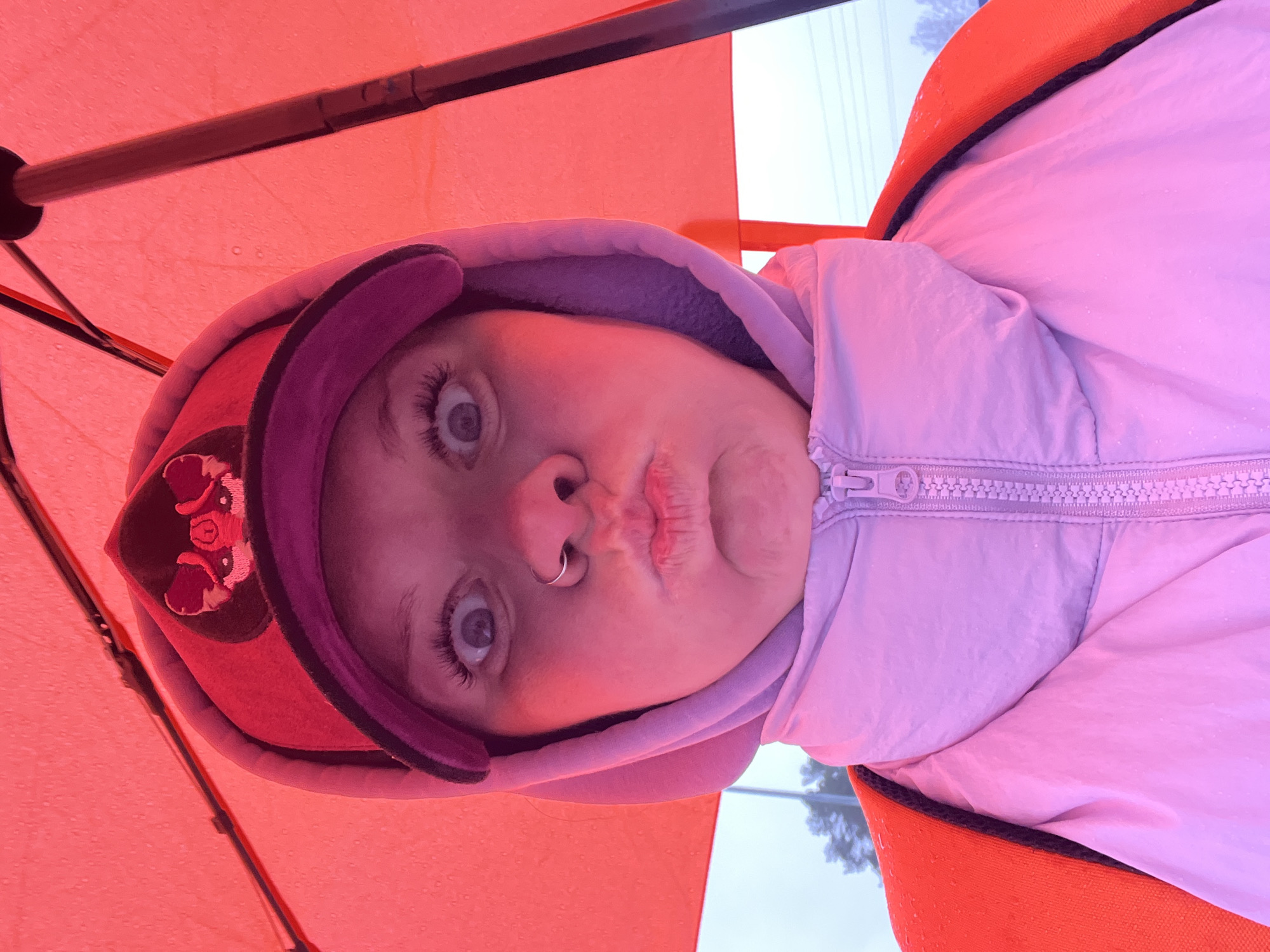 A close-up, low-angle selfie captures a person looking directly at the camera from underneath an orange-red umbrella. The person's face is centered, framed by a purple hooded jacket. Their eyes are wide open, and their lips are playfully pursed. A silver hoop nose ring is visible on their left nostril. On the left side of their head, part of a hat or a decorative element on the hood, possibly depicting a fox or similar animal in red and pink hues, can be seen.
The interior of the umbrella dominates the upper portion of the image, showing its orange-red fabric and a dark metal support rib. Small water droplets are visible on the umbrella's surface, indicating rain or recent precipitation. The person is dressed in a light purple/pink jacket with a visible zipper pull, layered under the purple hood. The background outside the umbrella is blurred and appears to be an overcast outdoor setting, with faint suggestions of distant trees or structures, consistent with a daytime scene in Chaikovsky, Russia. No visible text is present in the image. The overall mood conveyed is playful and candid, capturing a moment outdoors in inclement weather.