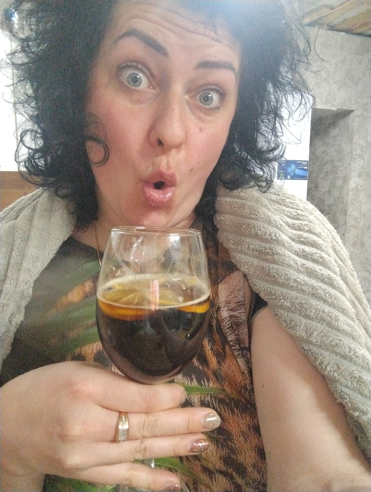 A woman with dark, curly hair is holding a glass of dark liquid with a slice of citrus fruit in it. She has a surprised expression, with wide eyes and pursed lips. She is wearing a patterned shirt, with a tan-colored towel draped over her shoulders. She has a gold ring with stones on her finger. The background appears to be an indoor setting, possibly a home or apartment. A TV screen can be seen in the background. The location is Grishanki, Russia.
