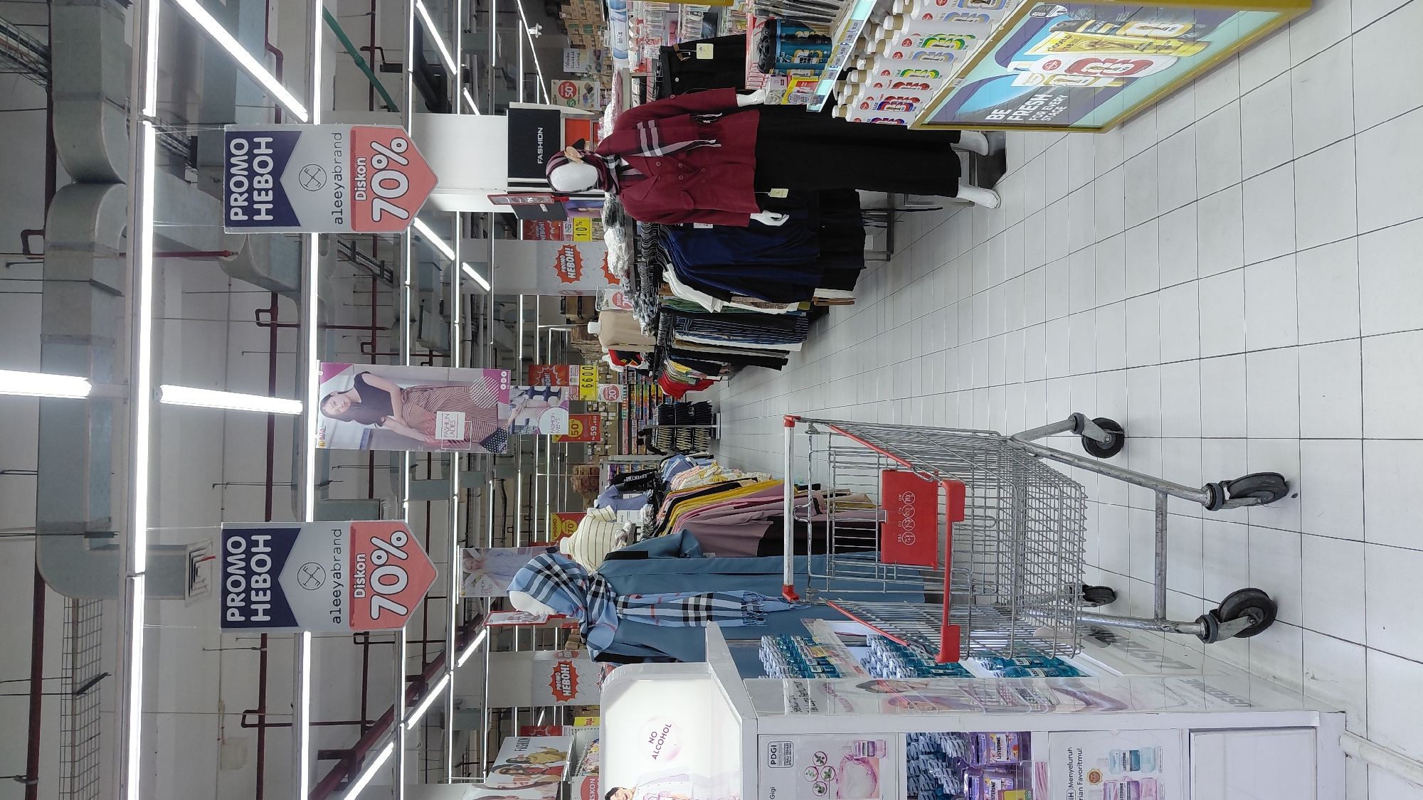 The image captures an interior view of a retail store, likely a department store or supermarket, in Purwokerto, Indonesia. The scene is brightly lit by fluorescent lights overhead, illuminating rows of clothing racks and display shelves. In the foreground, a silver shopping cart with a red basket sits on a white tiled floor. To the left, a display case holds various products, some labeled with "Listerine." Further back, mannequins dressed in clothing, including a blue outfit with a checkered scarf and a red jacket with a matching scarf, are positioned amongst the apparel racks. Several promotional banners are hung from the ceiling, advertising "PROMO HEBOH" and "Diskon 70%," indicating sales or special offers. The store appears to be well-stocked with a variety of clothing and other merchandise. The overall impression is one of a typical retail environment designed to attract shoppers.