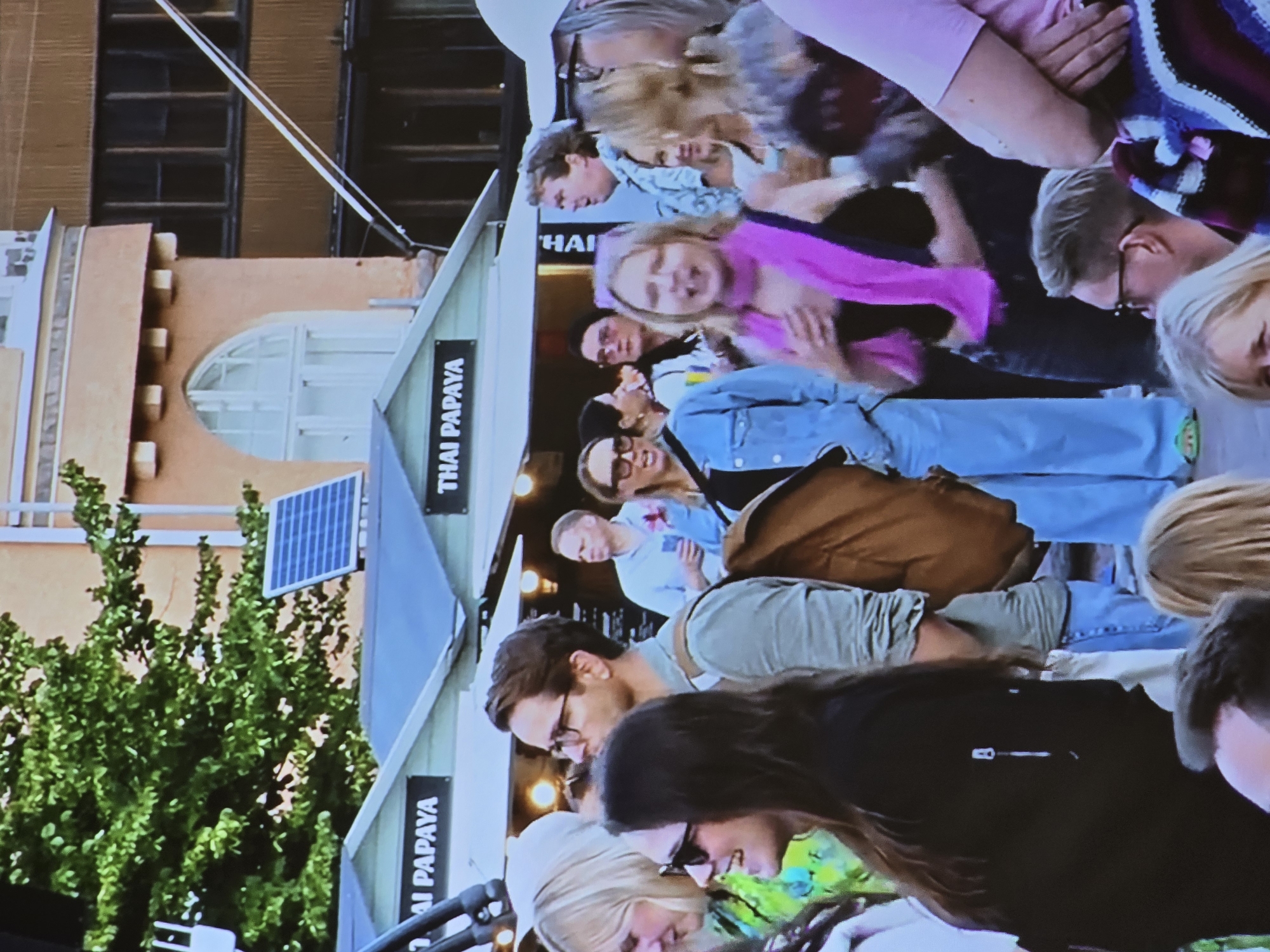 The image captures a bustling street scene in Helsinki, Finland, likely a market or festival area. Several people are visible, gathered around food stalls with signage reading "THAI PAPAYA." In the foreground, a woman with dark hair and sunglasses smiles, her face partially obscured by her hair. Next to her, a man with glasses and a light green shirt wears a backpack. Behind them, a crowd of people mill about, some holding drinks. One woman in the middle ground wears a bright pink scarf and appears to be speaking animatedly. In the background, a building with arched windows and decorative stonework suggests a historic urban setting. The overall atmosphere appears lively and social.