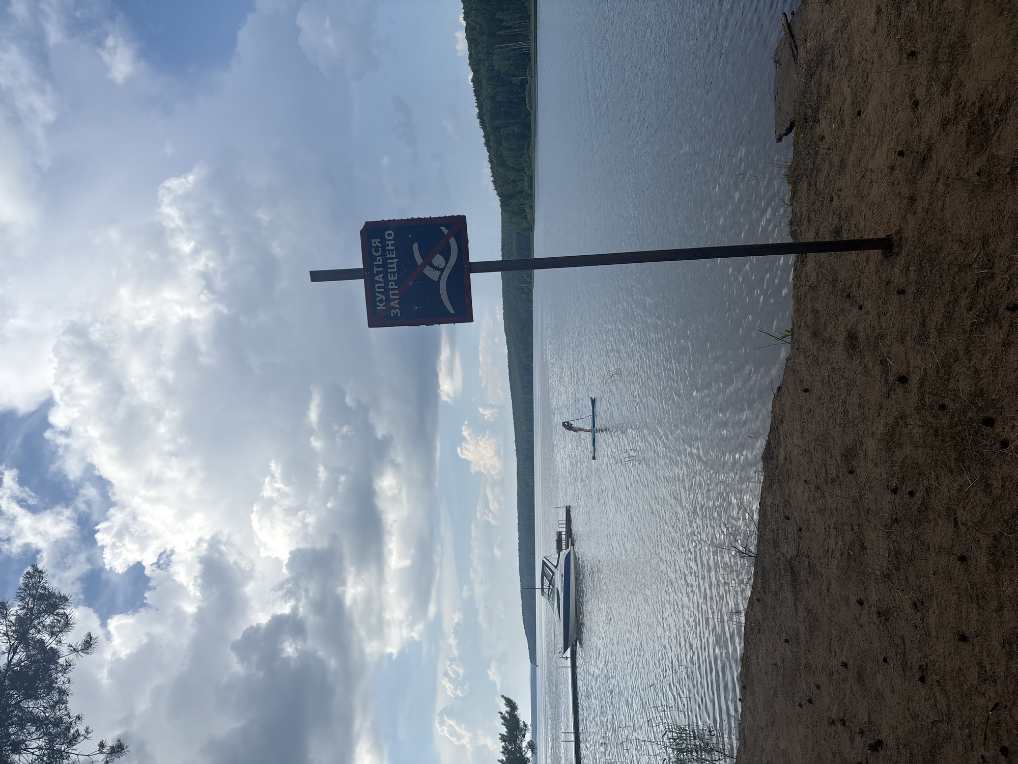 This image depicts a scenic view of the Kama River reservoir in Chaykovsky, Russia, on a cloudy day. The foreground features a sandy embankment with scattered pinecones and dry grass. A dark metal pole supports a square sign with a red border and a blue interior. The sign displays a white symbol of a person swimming with a diagonal red line across it, indicating "No Swimming."
In the middle ground, the calm, rippled surface of the water extends towards a wooden pier. A white motorboat is docked at the pier. Further out on the water, a person is visible on a stand-up paddleboard, seemingly unaware of or disregarding the "No Swimming" sign.
The background is dominated by the vast expanse of the water body, bordered by a dense, green treeline along the distant shore. The sky above is partly cloudy, with dramatic formations of white and grey clouds against patches of blue sky. The overall impression is one of a natural landscape with human recreational elements.