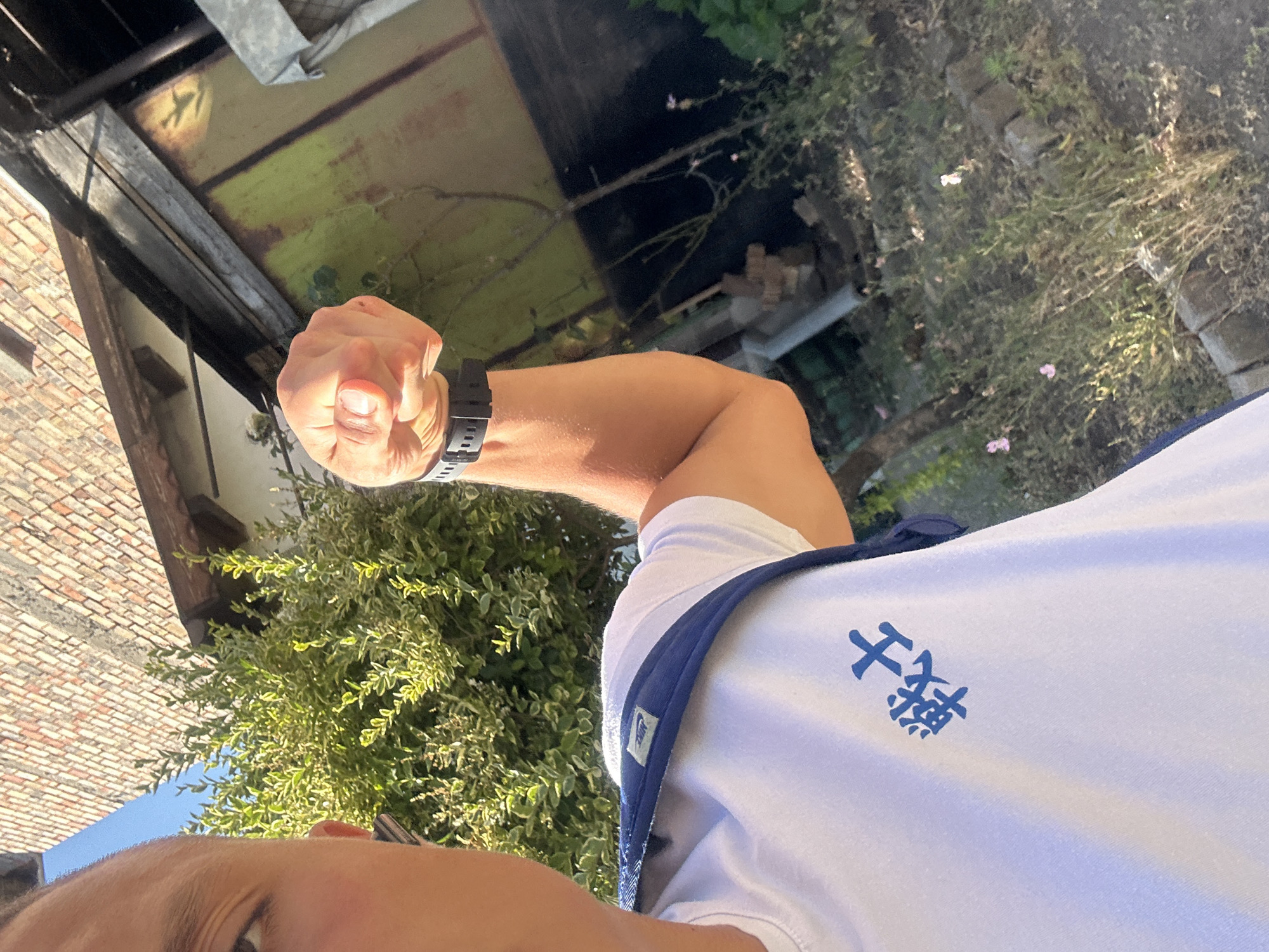 The image is a first-person perspective shot of a person's arm and upper torso, taken outdoors. The person is flexing their bicep, with a black watch on their wrist. They are wearing a white t-shirt with a blue trim and a blue symbol on the left chest area. The symbol appears to be two Chinese characters.
The background is a mix of an outdoor setting with greenery, including bushes and trees, and man-made structures. To the left, there is a brick wall and what appears to be part of a building with a weathered metal roof. Further back, there is more greenery and some scattered bricks on the ground. The lighting suggests it is daytime, likely sunny, casting shadows and highlighting textures. The overall scene appears to be a casual outdoor environment, possibly a garden or backyard, given the presence of foliage and building elements. There are no other people visible in the frame. The image is oriented vertically.