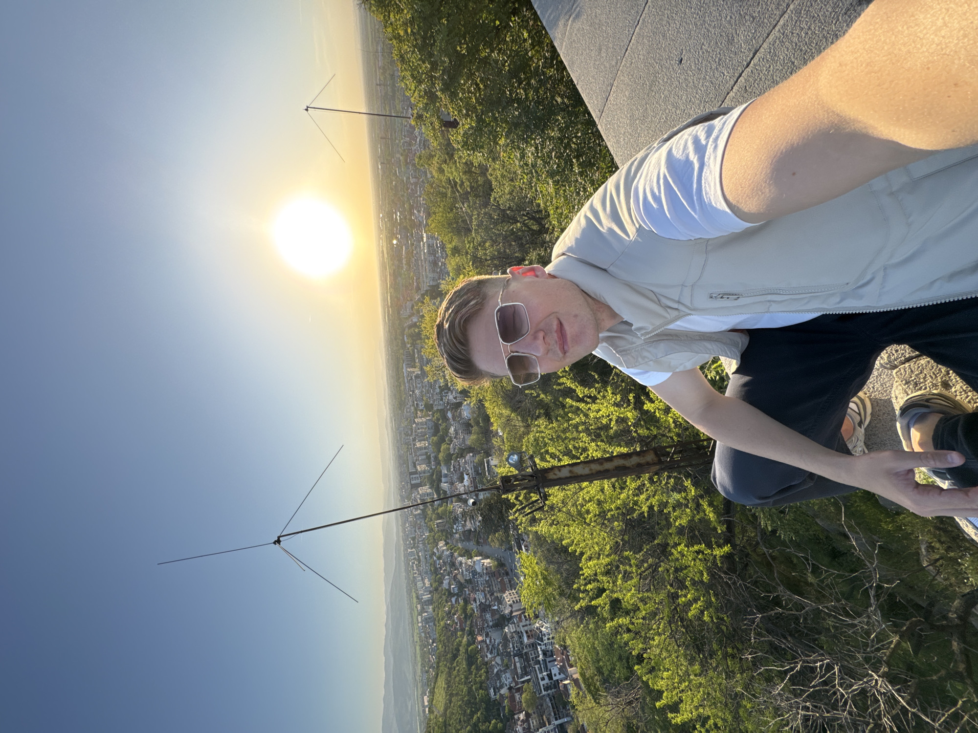 The image shows a selfie of a young man captured at what appears to be a high vantage point overlooking the city of Plovdiv, Bulgaria, during the golden hour. The man, positioned in the foreground and appearing to be crouching or kneeling, looks directly at the camera with a relaxed expression. He has light-colored hair and is wearing stylish sunglasses, a white t-shirt, a beige vest, dark pants, and sneakers.
Behind him, an expansive panorama unfolds. Below his position, lush green treetops give way to a sprawling cityscape with numerous buildings and structures, extending towards distant hills or mountains under a clear sky. The sun, a bright and intense orb, is low on the horizon, bathing the scene in a warm, golden-yellow light that gradually fades into a clear blue sky higher up. This strong backlighting creates a radiant glow around the sun.
Prominently featured in the middle ground are two metallic structures: one is a tall, slender mast with multiple wires extending outwards, resembling an antenna; the other is a thicker, more robust metallic beam. The overall setting suggests a scenic overlook or a rooftop, providing an impressive view of the city and natural landscape. The time of day is clearly either sunrise or sunset, indicated by the low sun and warm lighting conditions. No visible text is present in the image.