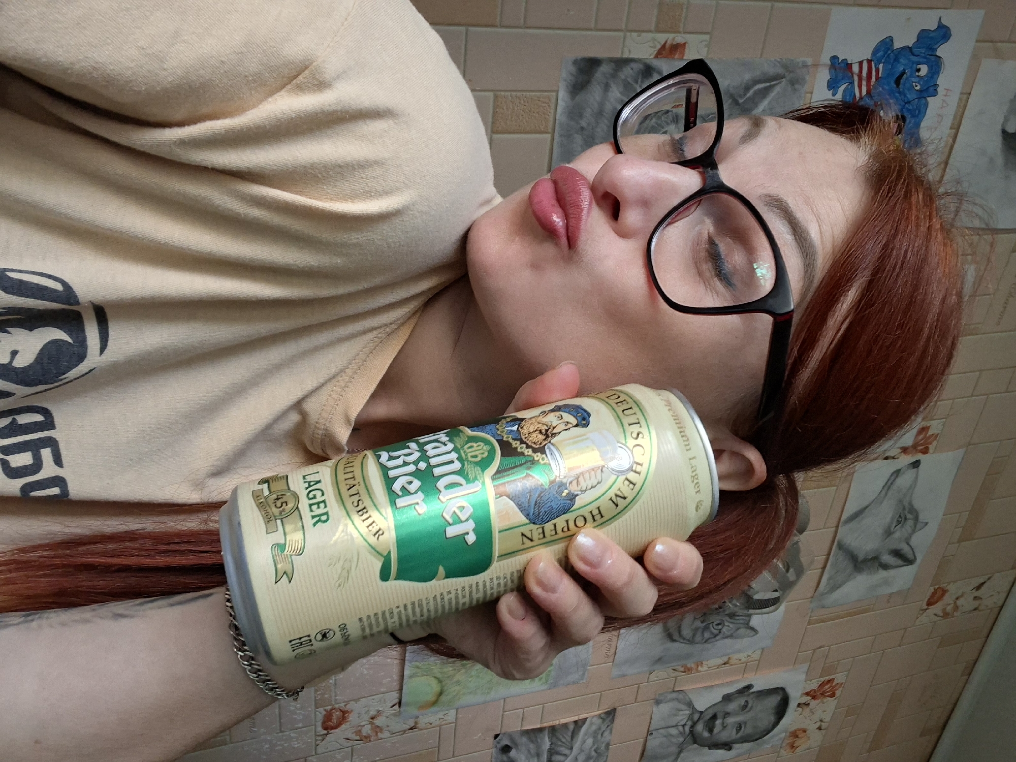 A close-up, indoor shot shows a woman with auburn hair pulled back into a ponytail, wearing black-rimmed glasses and a light peach-colored t-shirt. She is holding a can of "Brander Bier" lager, which has a label featuring an illustration of two men holding beer steins. Her right hand is around the can, and her left hand is partially visible. She is making a kissing face towards the camera, with her eyes closed. The background consists of a tiled wall adorned with several charcoal drawings, including a blue elephant, a fox, a cat, and a baby. The location is identified as Myadzyel, Belarus.