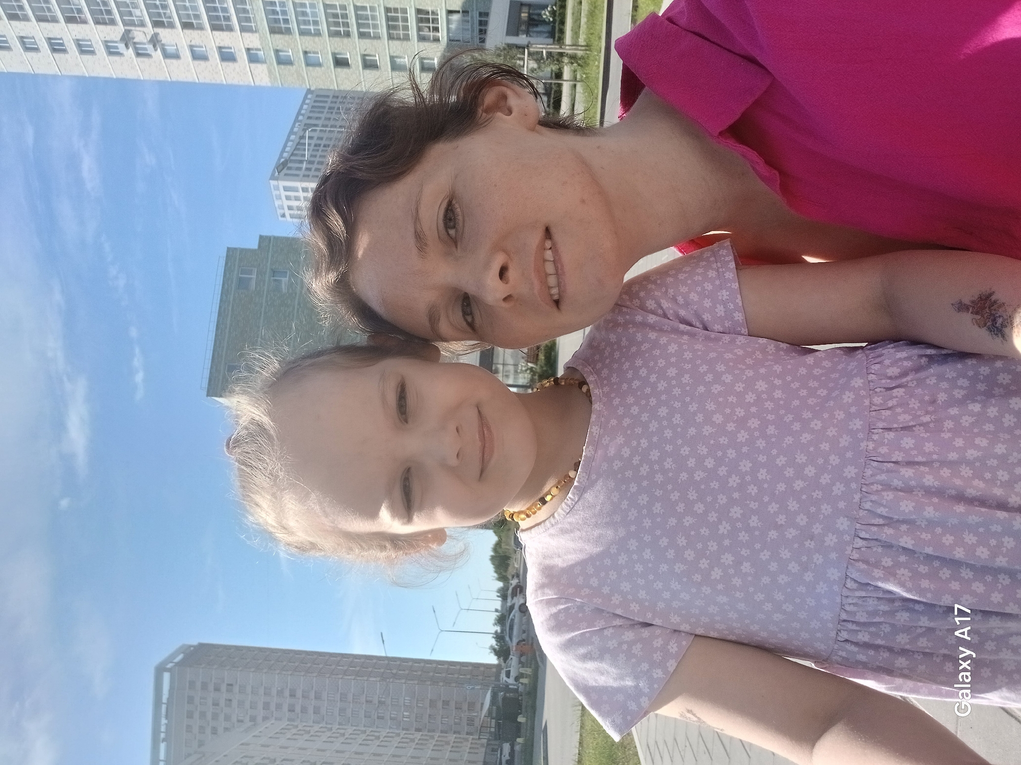 The image is an outdoor selfie of a fair-skinned woman and a young girl, seemingly a mother and daughter. The woman is wearing a pink shirt and has short brown hair. The young girl has light hair and is wearing a lavender dress with a floral pattern and an amber beaded necklace. The background shows a clear blue sky and several high-rise buildings. The location is in Yekaterinburg, Russia, based on the architectural style of the buildings. There's a street with streetlights visible in the background, and a sidewalk in the foreground. The image is slightly tilted, and the "Galaxy A17" watermark is visible in the lower right corner.