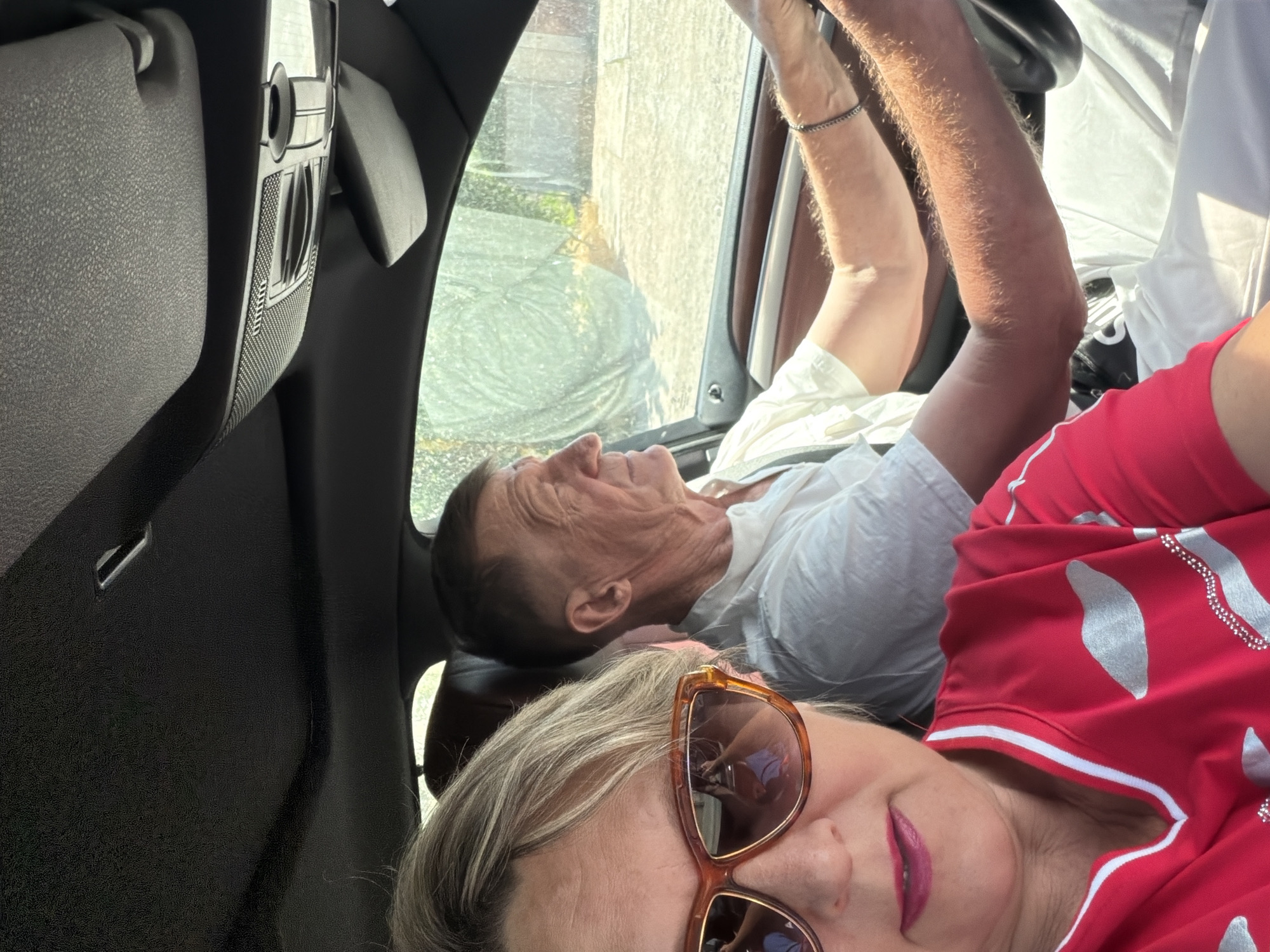 An over-the-shoulder selfie shows a woman with blonde hair and sunglasses in the foreground, smiling slightly. She is wearing a bright red shirt with silver embellishments. Behind her, a man with short brown hair is driving the car. He is wearing a white shirt. The car's interior is visible, including the dashboard and the roof lining. The exterior view through the car window shows sunlight filtering through some sort of netting or green foliage. The scene appears to be in a car, likely in a suburban or rural setting, based on the greenery and rustic fencing visible outside the window.