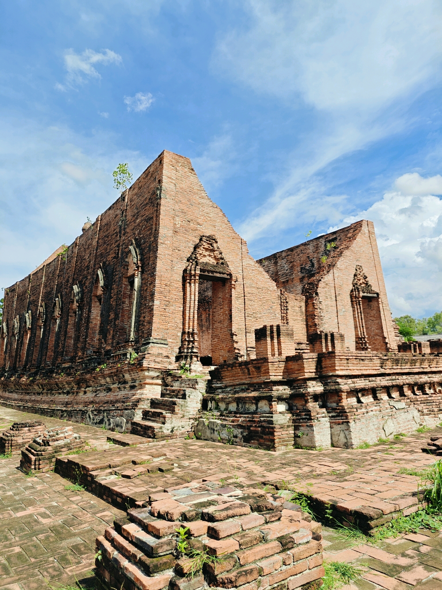An exterior, low-angle shot captures the ruins of a brick temple under a bright blue sky with scattered white clouds. The temple, constructed of weathered red bricks, features a curved facade with arched window-like indentations along its length. A prominent doorway, partially intact, is visible on the right side of the main structure. Steps and tiered brickwork lead up to the temple's base, and smaller brick ruins are scattered in the foreground. Green foliage sprouts from cracks in the bricks and among the surrounding pathways. The scene appears to be a historical site, possibly located in Ayothaya Town Municipality, Thailand, given the architectural style and brick construction commonly found in the region. No people are present in the image.