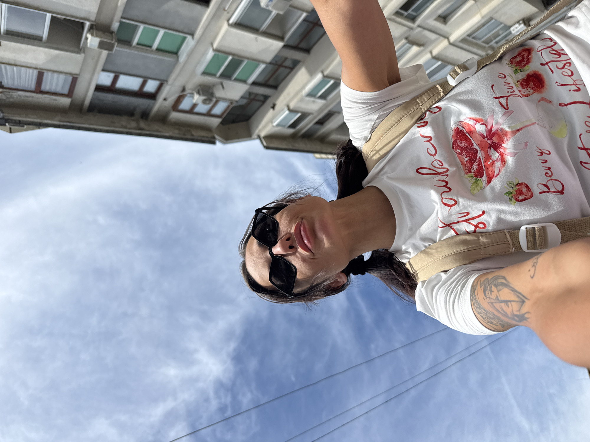 The media file shows a woman from the chest up, wearing a white t-shirt with a strawberry graphic and text. She is also wearing black sunglasses and has her hair in pigtails. The image is taken from a low angle, looking up towards the sky, with a building in the background. The sky is blue with some clouds, suggesting it is daytime. The woman appears to be outdoors, possibly in an urban environment given the building. The text on her t-shirt is in red script and appears to read "Strawberry Lifestyle" and "Berry Pure". The woman's expression is partially visible and she seems relaxed or amused. The overall impression is a casual, sunny day.