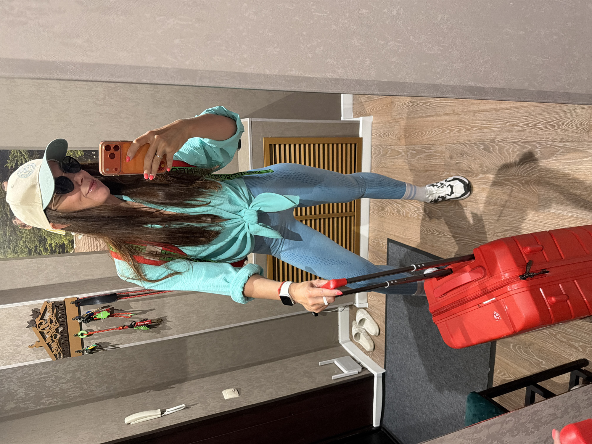 In a hallway in Birobidzhan, Russia, a woman stands in front of a mirror, taking a selfie. She is wearing a teal cropped blouse tied at the waist, light blue athletic leggings, a cream and teal baseball cap, and sunglasses. She also has a red backpack on and is holding the handle of a red rolling suitcase with her right hand. Her left hand, adorned with a white ring and a red bracelet, is holding her orange smartphone, which is pointed at the mirror. The hallway features textured gray walls, a wooden floor, a dark brown door, a key rack with several keys hanging on it, a shoe rack with slippers, and a small stool with a dark green cushion.
