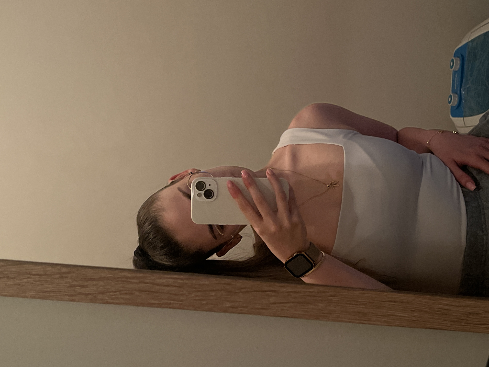 The image displays a mirror selfie, rotated 90 degrees counter-clockwise. The central subject is a person, whose upper body is visible in the reflection. They are wearing a plain white or light-colored sleeveless top and glasses with thin frames. Their dark hair is partly visible on the left side of the frame, possibly tied back.
Around their neck, they wear a delicate golden necklace with a small charm. Their left wrist sports an Apple Watch with a gold-toned case and band, along with a subtle bracelet. The person's right hand holds a white smartphone, prominently featuring a triple-lens camera system, which is being used to take the picture. Their fingernails are painted a light, neutral shade. The person's face is largely obscured by the phone.
The setting appears to be indoors. The reflection shows a plain, light beige wall in the background. A light brown wooden ledge or shelf runs horizontally across the lower part of the frame, indicating a surface below the mirror. In the upper right corner, a portion of a blue and white object, possibly an appliance or storage unit with circular elements, is partially visible.
The activity depicted is the taking of a mirror selfie. The lighting is soft and even, consistent with indoor conditions. No specific time of day or outdoor weather elements are apparent. Small, unidentifiable text is faintly visible on the blue and white object in the background.