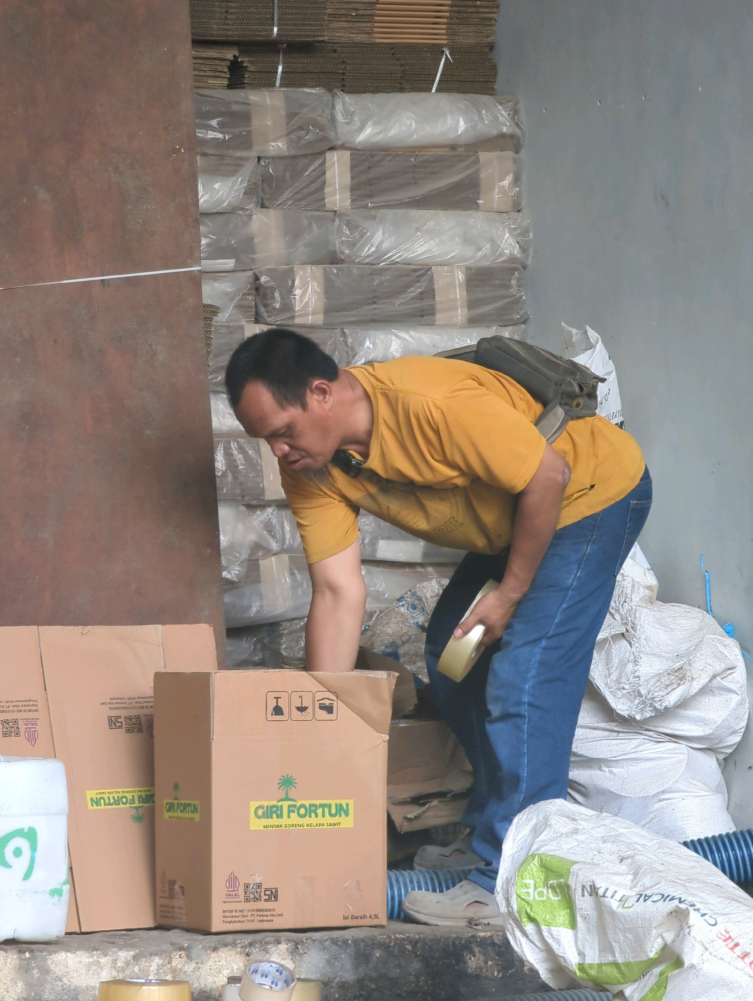 A man in a yellow t-shirt and blue jeans is seen from the back in a warehouse setting in Batu Belaman, Indonesia. He is bent over, holding a roll of tape in his right hand and appears to be working with cardboard boxes stacked behind him. The boxes are wrapped in clear plastic, suggesting they are being prepared for storage or shipping. Piles of white sacks and a blue hose are also visible in the background, indicating a storage or industrial environment. The lighting suggests it is daytime, with diffused light coming from an unseen source. The man's posture and the presence of packing materials suggest an ongoing activity related to logistics or inventory management within the warehouse.