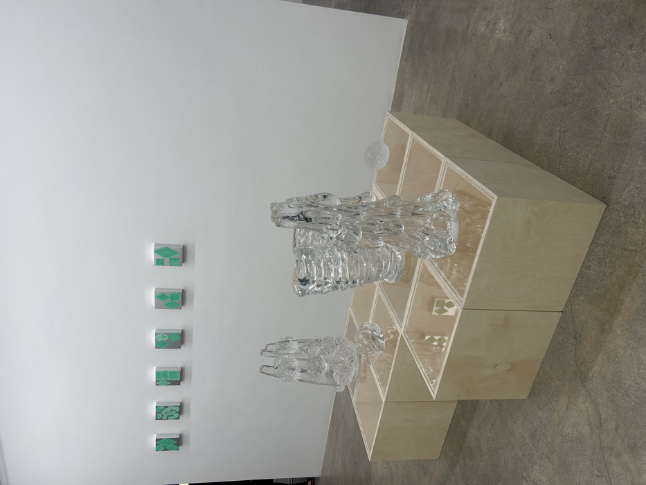 The image displays an art installation. A white wall features a vertical series of six small square artworks with green geometric patterns. To the right stands a light-colored, tiered, geometric shelving unit, showcasing two clear glass sculptures with complex, flowing forms. A smaller, translucent sphere sits atop one section of the shelving. The background includes a plain concrete floor.