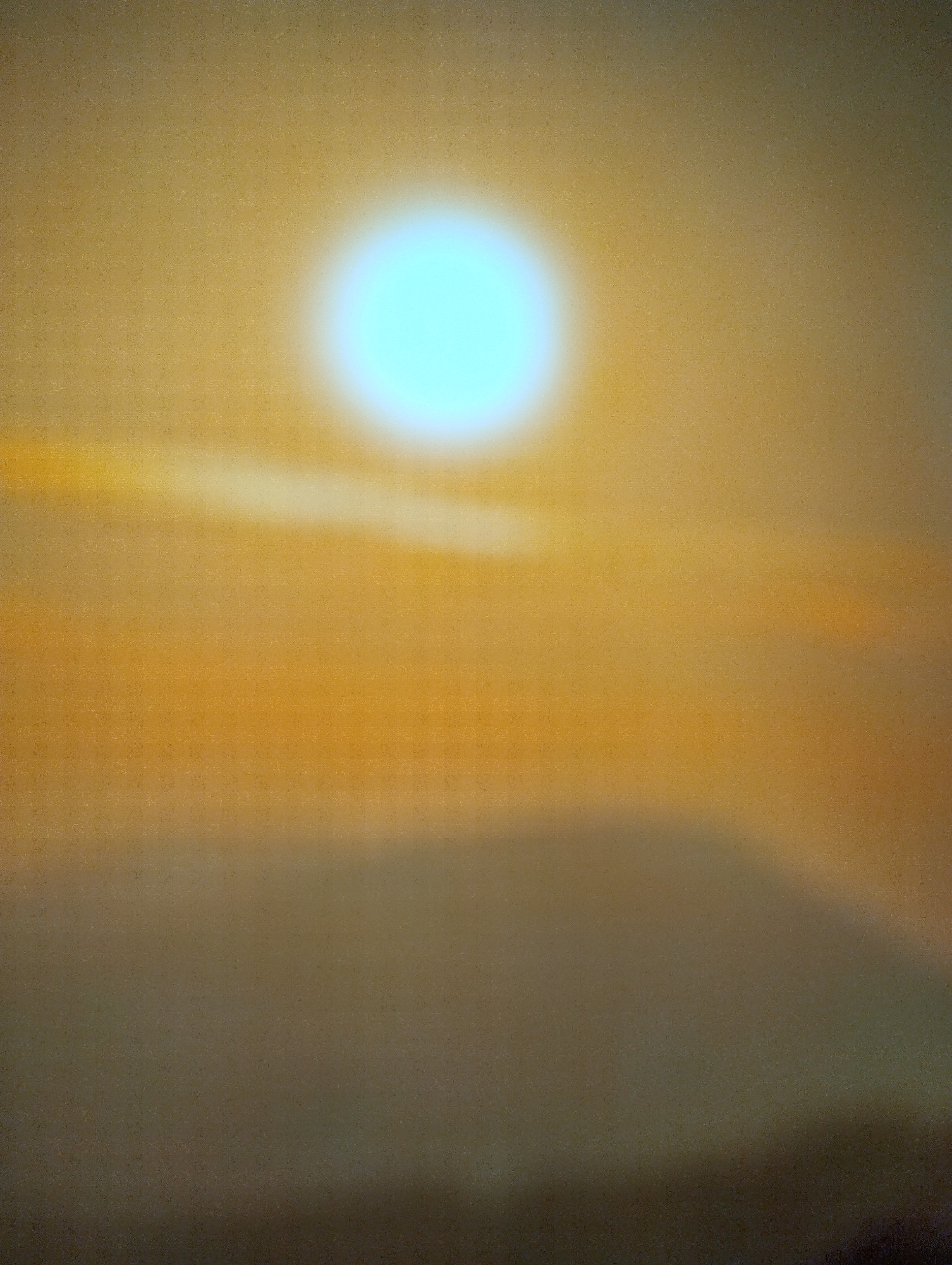 The image presents a blurred scene in Martil, Morocco. A bright, out-of-focus, light blue orb dominates the upper portion of the frame. Hues of yellow, orange, and brown blend together, creating an abstract landscape that is predominantly blurred. The bottom section displays darker tones, suggesting either a blurred surface or land. The overall blur obscures any specific details about objects, people, or actions.