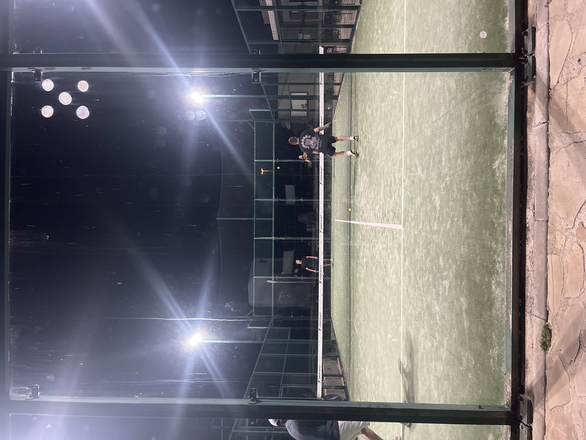 The image shows a padel tennis match in progress at night in el Poble Nou de Benitatxell / Benitachell, Spain. Two players are on the court, one near the net about to return a shot, and the other closer to the back fence. The padel court is surrounded by metal fencing and glass panels, which are illuminated by overhead lights. The ground outside the court is paved with stone. The sky is dark, indicating it is nighttime. The setting suggests an outdoor sports facility within the mentioned Spanish city.