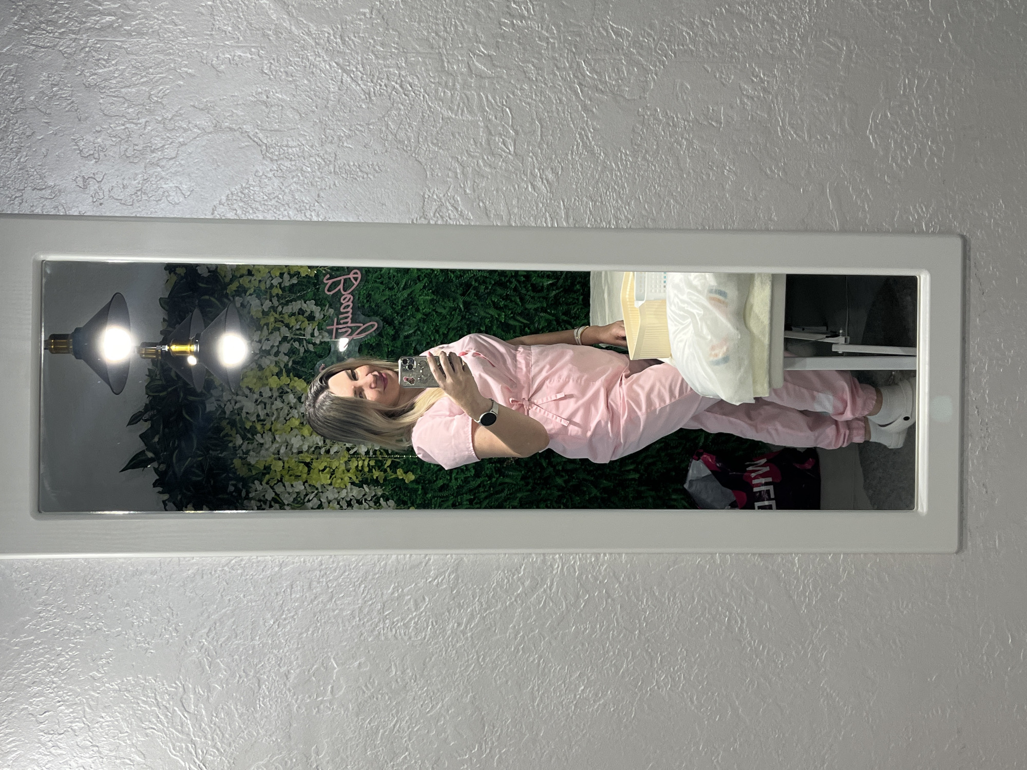 The image captures a full-length mirror selfie of a woman in an indoor setting, likely in Sochi, Russia. The woman, positioned centrally, is reflected in a rectangular mirror with a white frame. She is wearing light pink scrubs or a uniform, a white smartwatch on her left wrist, and white shoes (possibly Crocs). She is holding a smartphone in her right hand, taking the photograph, and has a slight smile on her face. Her blonde hair is styled down.
The background reflected in the mirror features a vibrant green wall covered in artificial foliage, giving the impression of a plant wall. Two industrial-style pendant lights with exposed bulbs hang down, casting bright light. To the left of the woman's head, a pink neon sign is visible, displaying the word "Beauty" in a stylized, cursive font, possibly with a small decorative element like eyelashes or a lipstick.
To the right of the woman, a counter or shelf holds several items, including white plastic bags and a yellow container. On the floor, near her feet, a black bag with "MFE" or "BME" partially visible in white text is reflected. The wall surrounding the mirror has a textured, stucco-like finish, painted in a light, neutral color. The overall setting appears to be a modern commercial space, such as a beauty salon, spa, or clinic. The time of day is not discernible, as the scene is artificially lit.
