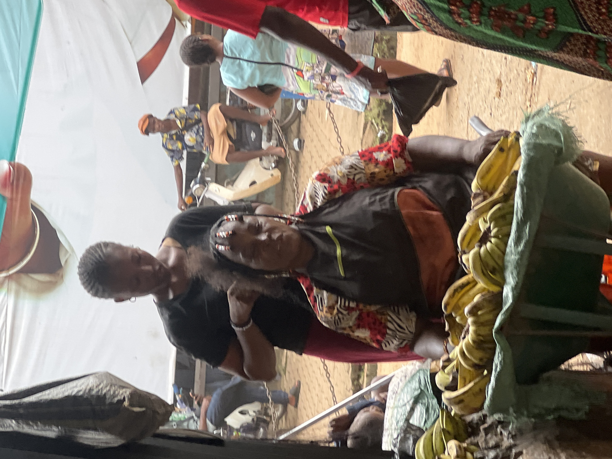 This image depicts a bustling outdoor market scene in Nkpor, Nigeria. In the foreground, a woman with braids adorned with beads is being attended to by another woman. The woman receiving the service wears a colorful patterned shirt and an apron. Next to them, a cart is filled with ripe yellow bananas, suggesting a fruit stall. In the background, other individuals are visible, including a person sitting on a motorbike wearing an orange cap and a patterned shirt. The environment appears to be a typical marketplace with various people engaged in their daily activities.