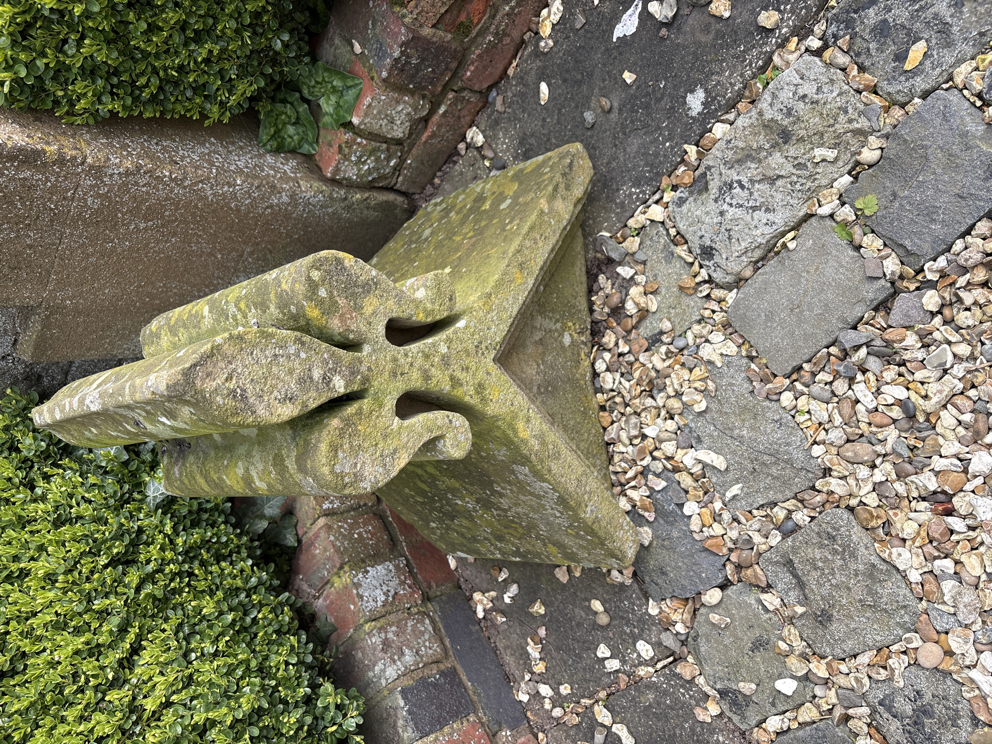 The scene presents an exterior view at ground level, likely within a garden or yard in Pulverbatch, United Kingdom. A weathered stone object, possibly a decorative architectural fragment, is centrally positioned. It's greenish-grey and displays signs of age and weathering, with lichen or moss covering portions of its surface. The object is angular with geometric cut-outs. The object sits at the edge of a brick wall and adjacent to a stone pavement area. The pavement is composed of grey stone blocks interspersed with small gravel stones. Adjacent to the stone object and brick wall is a dense, green bush, possibly boxwood. The ground cover includes ivy, adding further textures.