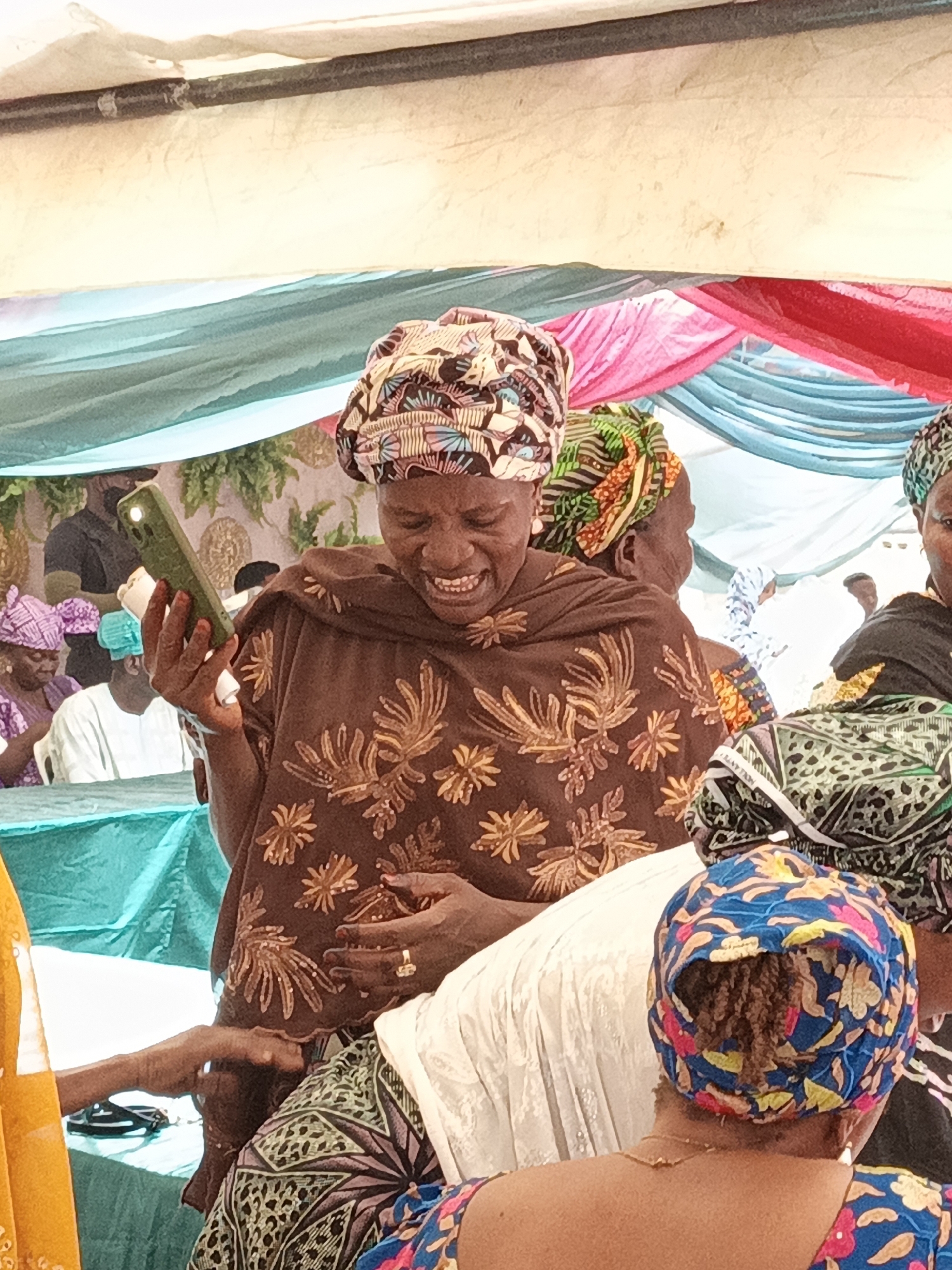 The image captures a lively social gathering, likely an event or celebration, in Sabon Gari West, Nigeria, during daylight hours. The central focus is a woman in a brown garment adorned with gold/light brown floral patterns and a vibrant, multi-patterned head wrap. She is smiling broadly, almost laughing, as she looks down at a green smartphone held in her left hand, which also clutches a rolled white object with blue markings. Her expression conveys joy or amusement.
Around her, several other women are visible, also dressed in patterned traditional attire and head wraps, some partially obscured. One woman's back is visible in the foreground wearing a blue head wrap with colorful floral designs. In the background, draped fabrics in shades of light blue, pink, and red serve as festive decorations, possibly lining a tent or indoor venue. A teal/turquoise tablecloth is visible in the lower left, where other attendees are seated. A man in a dark shirt is partially visible behind the central woman, also seated. The overall atmosphere appears joyful and social.