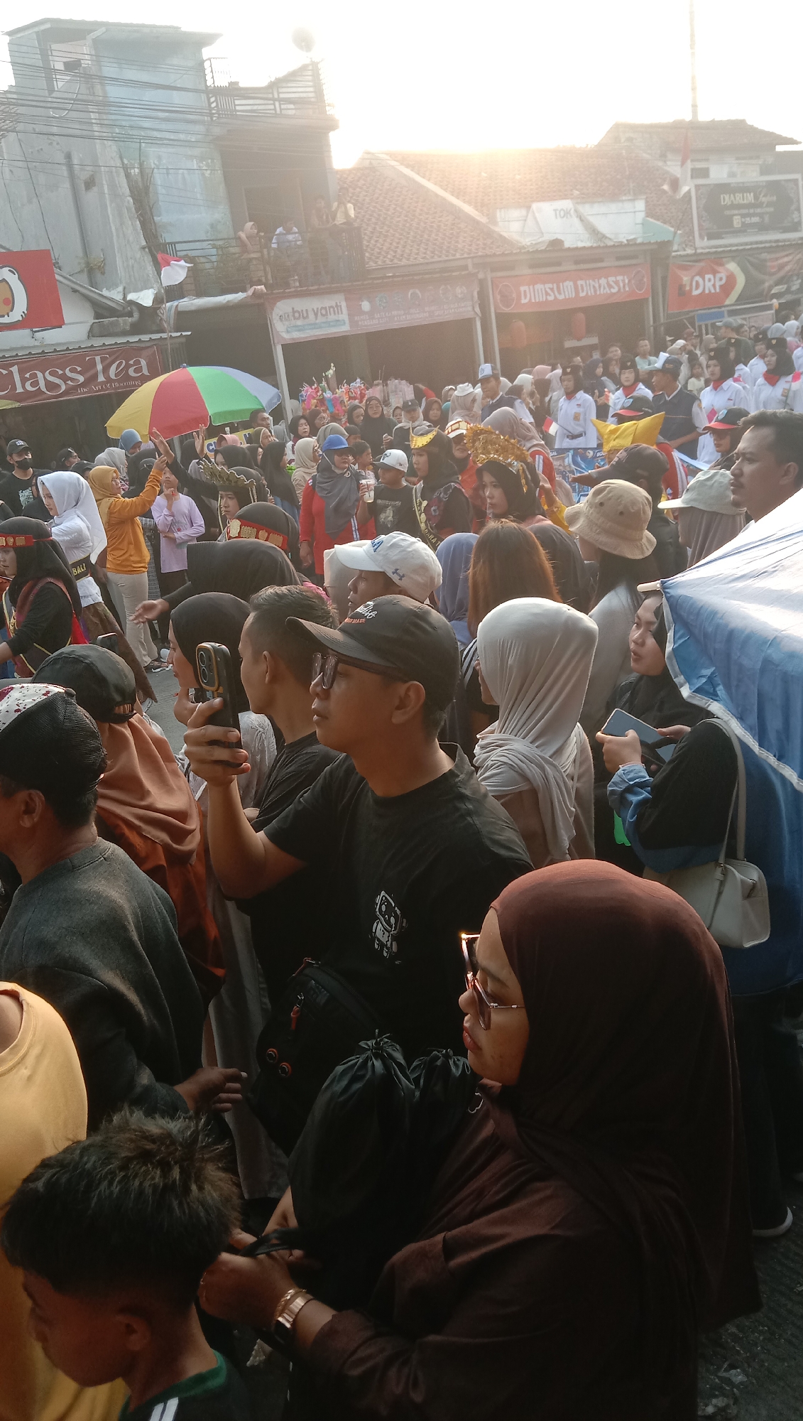 The image captures a street scene in Jatisawit, Indonesia, during what appears to be a public gathering or event, possibly a parade or festival. The atmosphere is crowded, with many people densely packed together. Buildings line the street, some with visible signage indicating businesses. The sun is low in the sky, casting a warm glow and creating lens flare in some areas.
In the foreground, a man wearing a black baseball cap and glasses is holding a smartphone, seemingly taking a photo or video. To his right, a woman in a brown hijab is also looking to the side, and further to her right, another woman in a white head covering is holding a phone. A young boy with spiky hair is visible on the far left.
The crowd is diverse, with many individuals wearing casual clothing, while some are adorned with festive attire, including headwear and costumes, suggesting a celebratory event. There are also various objects present, such as umbrellas, bags, and possibly stalls or decorations associated with the event. The overall impression is one of active public participation in an outdoor event within an urban environment.