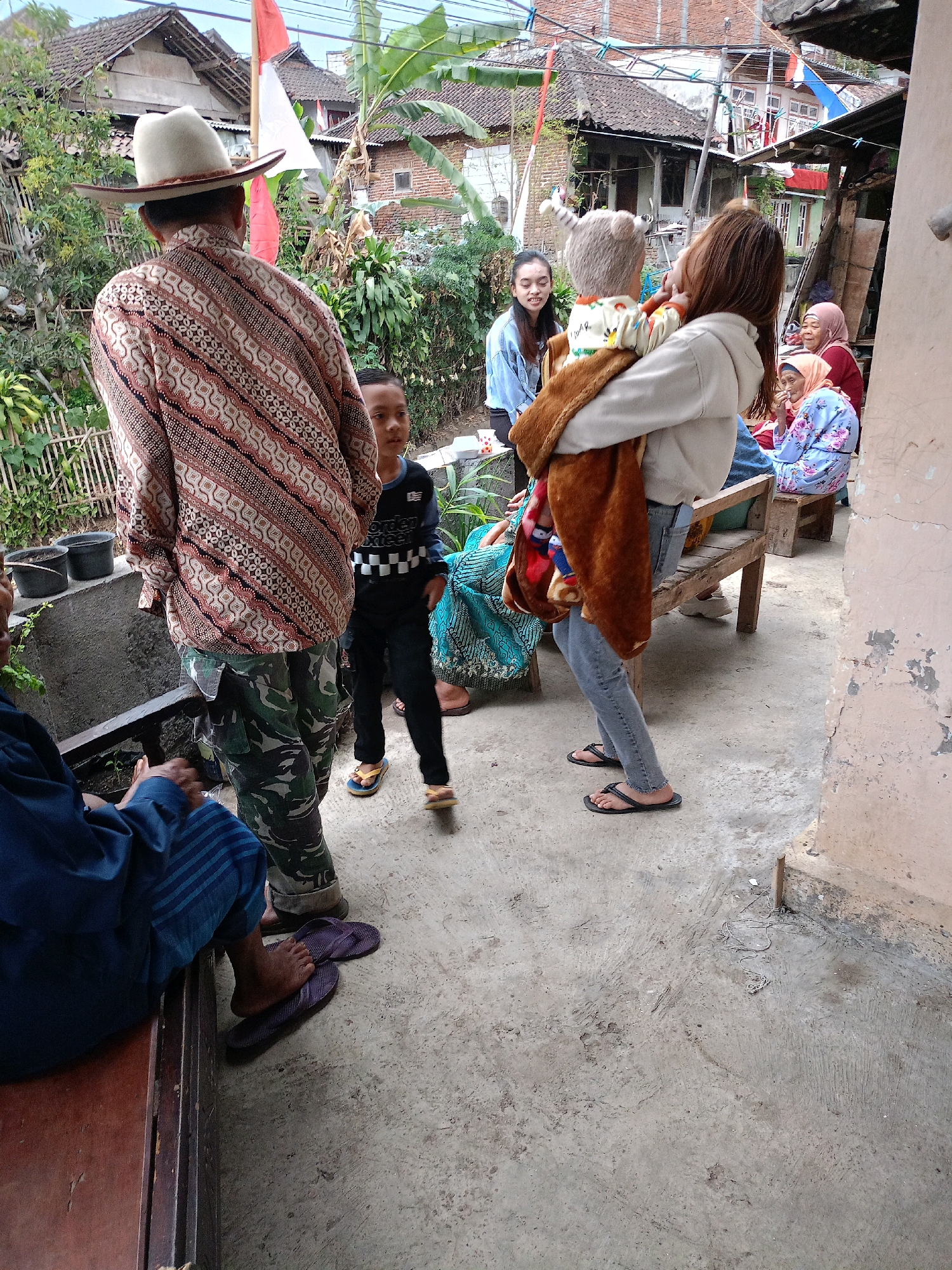 In Batu, Indonesia, a scene unfolds on a concrete patio with a backdrop of houses and greenery. A man wearing a wide-brimmed hat and a batik patterned shirt stands facing away from the camera, his camouflage pants a striking contrast. To his right, a young boy in black pants and a patterned shirt walks away, his yellow flip-flops visible. In the mid-ground, a woman with long brown hair cradles a baby wrapped in a brown blanket. Another young woman in a blue denim jacket looks on, her expression curious. Further back, two older women are seated on a wooden bench, one wearing a pink headscarf and the other a floral blue patterned headscarf. The environment is cluttered with potted plants, a wooden bench, and discarded items. The overall impression is of a casual gathering in a residential area, possibly a courtyard or a front yard.