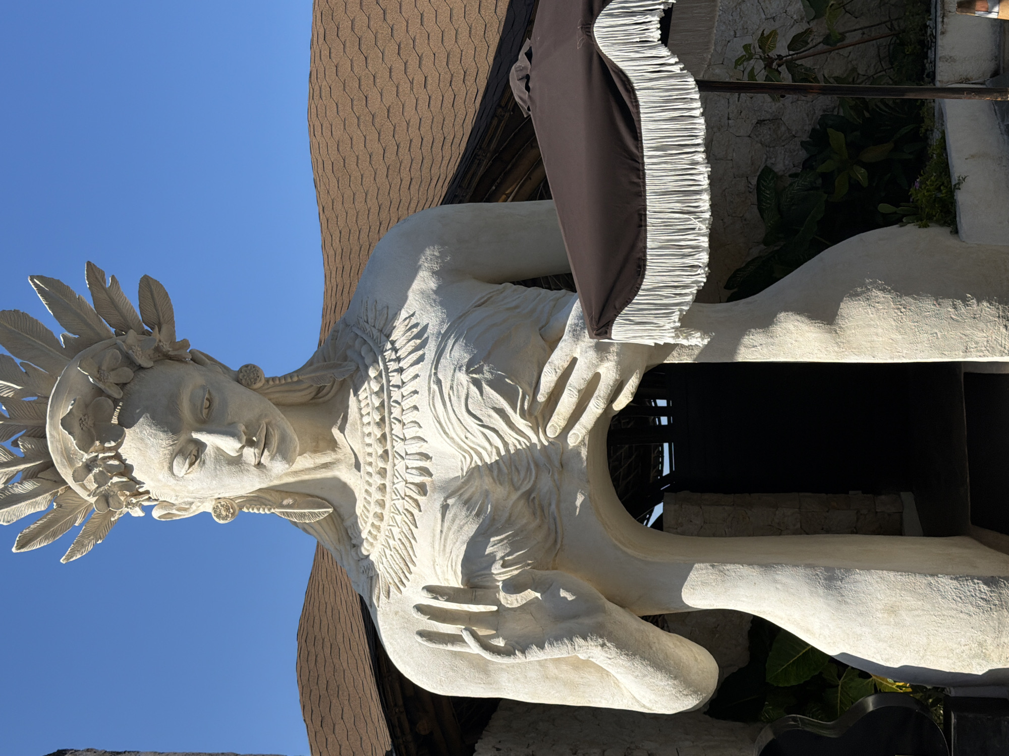 The image depicts a weathered, white-colored sculpture of a woman with a head adorned with feathers and floral motifs. She has her hands placed across her chest. The sculpture appears to be outside, under a clear blue sky, adjacent to a building with a brown-colored, shingled roof. A fringed awning or canopy, brown and white, partially obscures the sculpture. The setting is Ungasan, Indonesia.