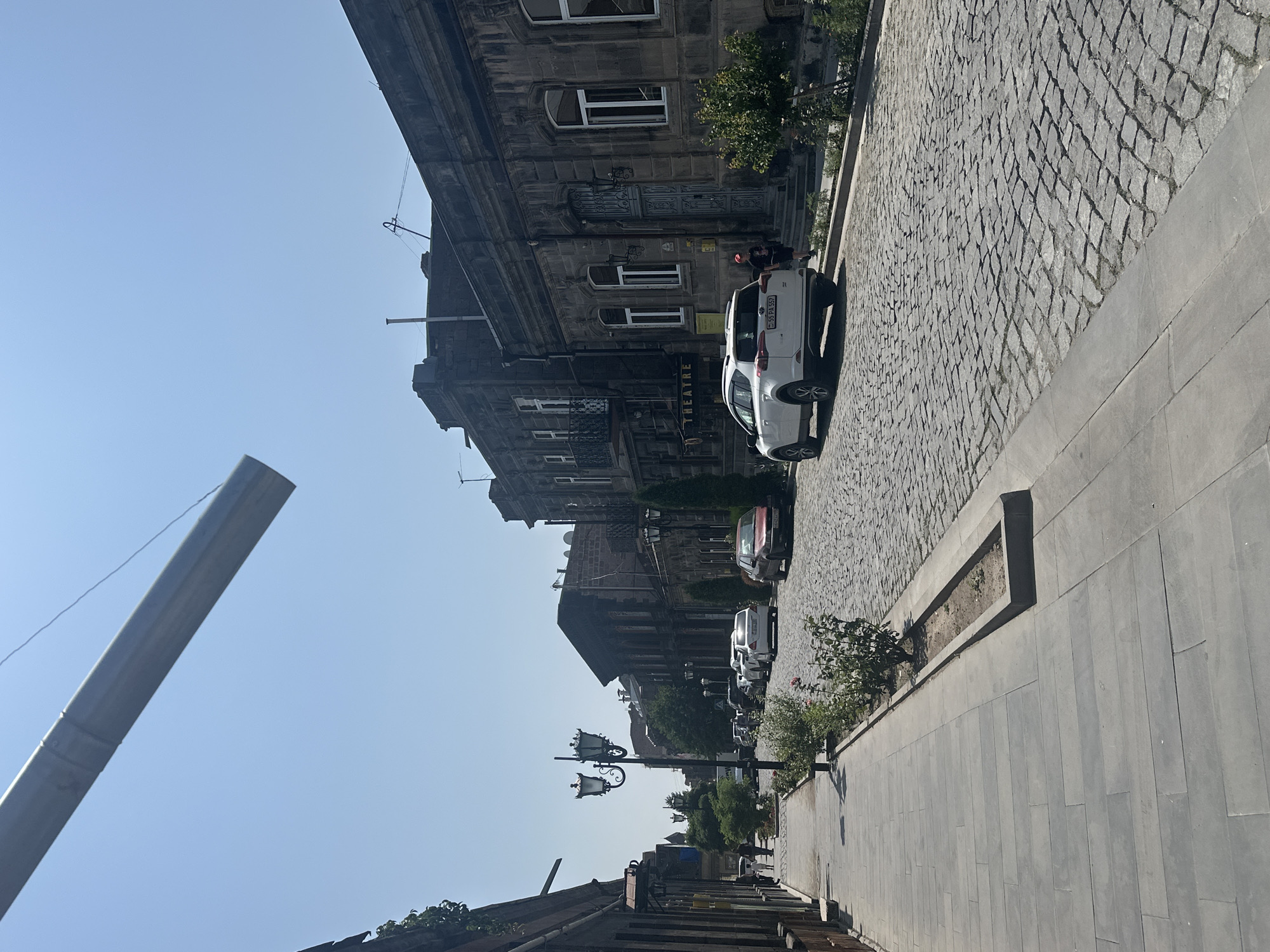 A clear, sunny day in Gyumri, Armenia, is captured in this vertical, eye-level, medium-shot photograph. The scene depicts a street lined with historic stone buildings, a cobblestone road, and a paved sidewalk. The architecture suggests an older part of the city, with ornate stone facades and balconies. A white SUV is parked on the right side of the cobblestone street, partially obscuring a red sedan parked behind it. A few pedestrians can be seen walking on the sidewalk on the left side of the street. A decorative lamppost stands on the left side of the street, casting a shadow on the sidewalk. In the foreground, a gray pipe extends from the upper left corner of the frame, obscuring a portion of the blue sky. The street is generally clean, with some greenery planted in flower beds along the sidewalk. The overall impression is of a well-preserved historic urban environment.