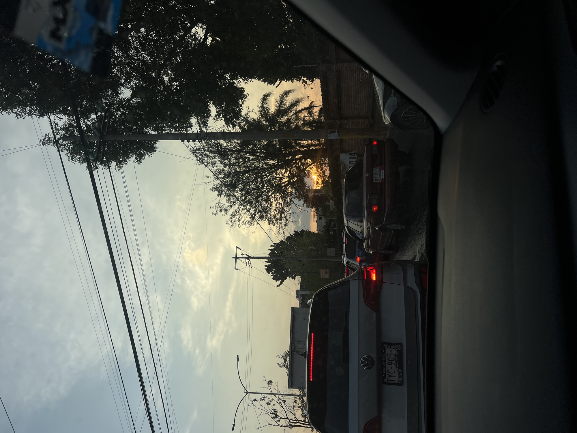 The image, taken from inside a car, shows a street in Puebla, Mexico, likely during a sunset or sunrise. The view is through the windshield, with the car stopped in traffic. Visible in the foreground are the rear ends of several vehicles. The vehicle closest to the camera is a silver Volkswagen Taigun with the license plate TYC-905-B from Puebla, Mexico. Ahead of it is a red Mazda Tsuru with license plate TPP-450-B. Electrical wires and poles line the street, and trees are visible on both sides. A palm tree is visible with the sun appearing behind it, casting a warm glow on the scene. The sky is bright, suggesting the time of day. A brick wall and the side of a building are present on the right.