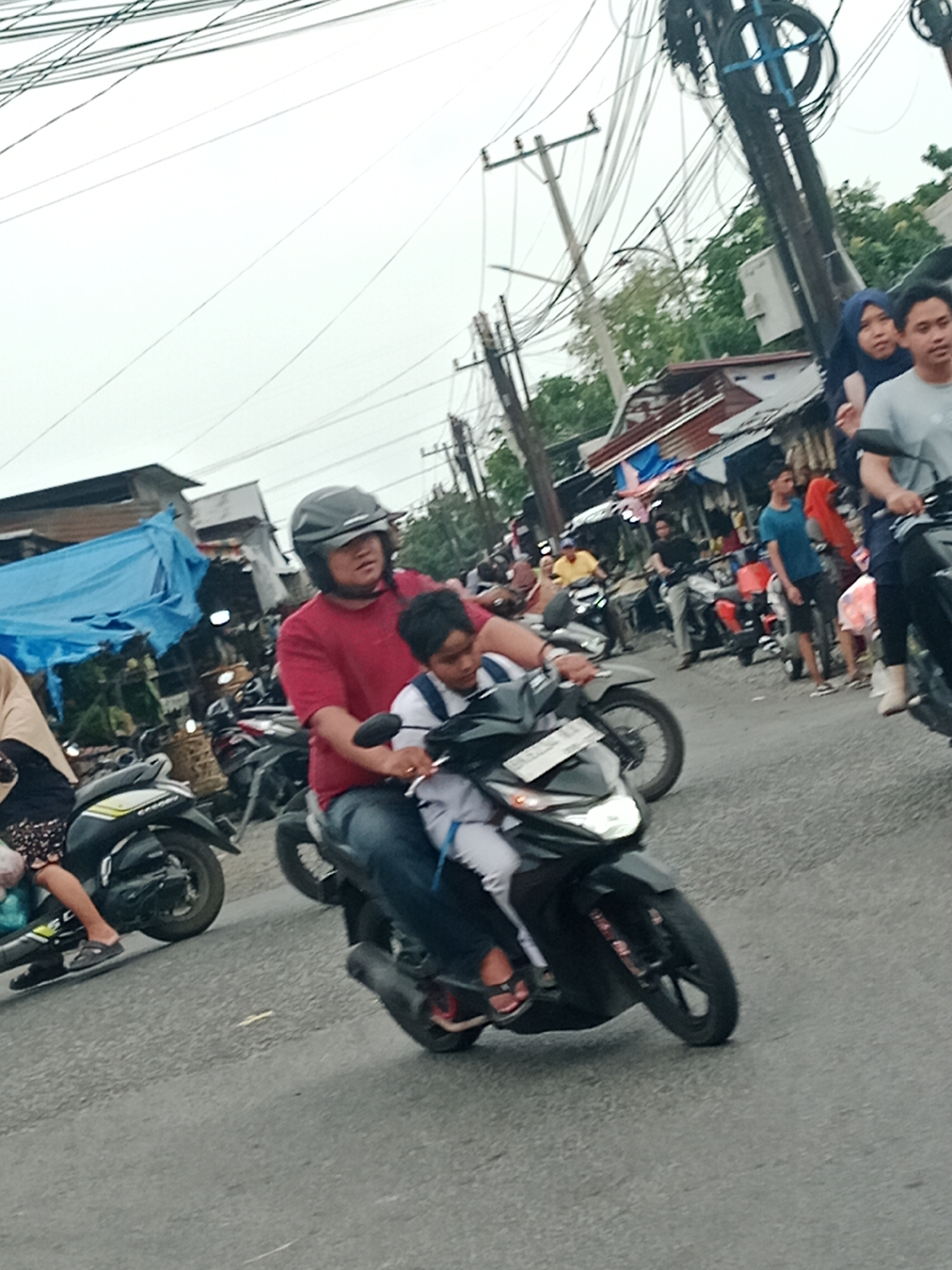 In the City of Binjai, Indonesia, this image captures a street scene with multiple motorcycles and pedestrians. In the foreground, a man wearing a red shirt and a grey helmet is riding a black motorcycle, with a younger individual in a white shirt as the passenger. The rider's left hand is on the handlebars, and the passenger is looking forward. To the left, another motorcycle is partially visible, with a person wearing a hijab and patterned skirt on the passenger seat. In the background, several other motorcycles and people are present, suggesting a busy urban environment. Structures resembling market stalls or small shops line the side of the road, with tarps and corrugated metal roofs. Electrical wires crisscross overhead, indicating utility infrastructure. The road surface is asphalt. The overall atmosphere appears to be one of daily commerce and transportation.