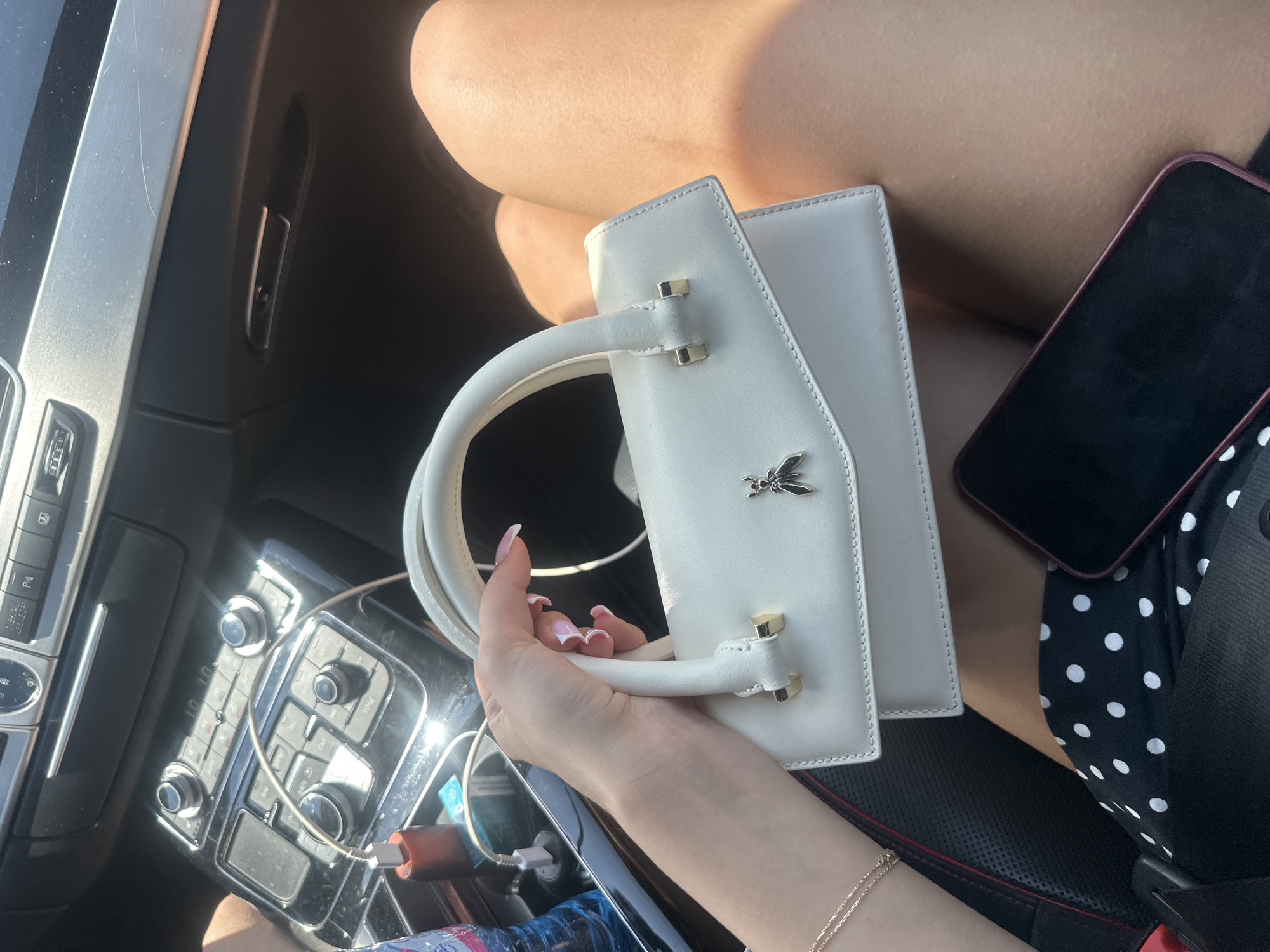 The image shows a close-up of a white handbag with a gold bee emblem, held by a person with manicured fingernails. The handbag is placed on the lap of a person wearing a polka dot skirt and a seatbelt. In the background, the dashboard of a car is visible, with buttons and a charging cable plugged into a port. The scene appears to be inside a car, likely in the city of Polikraishte, Bulgaria, as suggested by the context. The lighting suggests it is daytime.