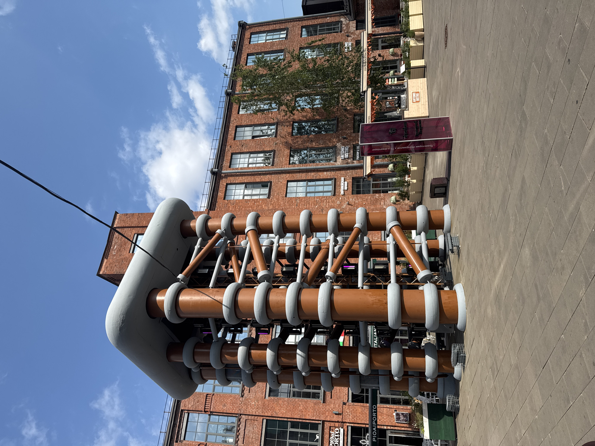 The image captures an outdoor view of a unique art installation, likely a Tesla coil sculpture, situated in the courtyard of a brick building complex. The sculpture is large and intricate, with multiple copper-colored vertical columns adorned with grey, ring-like insulators. Orange pipes crisscross between the columns, connecting them and contributing to the complex structure. The sculpture is grounded by square wire cages.
Behind the sculpture, a red brick building with numerous large windows stands prominently. The windows are multi-paned and reflect the sky and surrounding environment. The building appears to be a repurposed industrial structure, likely part of a larger complex known for its artistic or cultural significance. Signs for "PIOAIPORTO," "ITALIAN," and "ANTONIO BISTRO" are visible on the building's facade, suggesting a commercial or gastronomic area.
The courtyard itself is paved with light-colored square tiles, creating a clean and modern appearance. There are a few small green planters and seating areas visible, indicating this is a public space designed for visitors. The sky is a vibrant blue with scattered white clouds, and the sunlight suggests it is daytime.
The art installation and the surrounding architecture strongly suggest the location is **Sevkabel Port in Saint Petersburg, Russia**. Sevkabel Port is a well-known cultural and entertainment hub built on the former territory of the "Sevkabel" cable plant, and it features many industrial-chic architectural elements and public art installations. The style of the brick buildings and the presence of contemporary art are characteristic of this space.