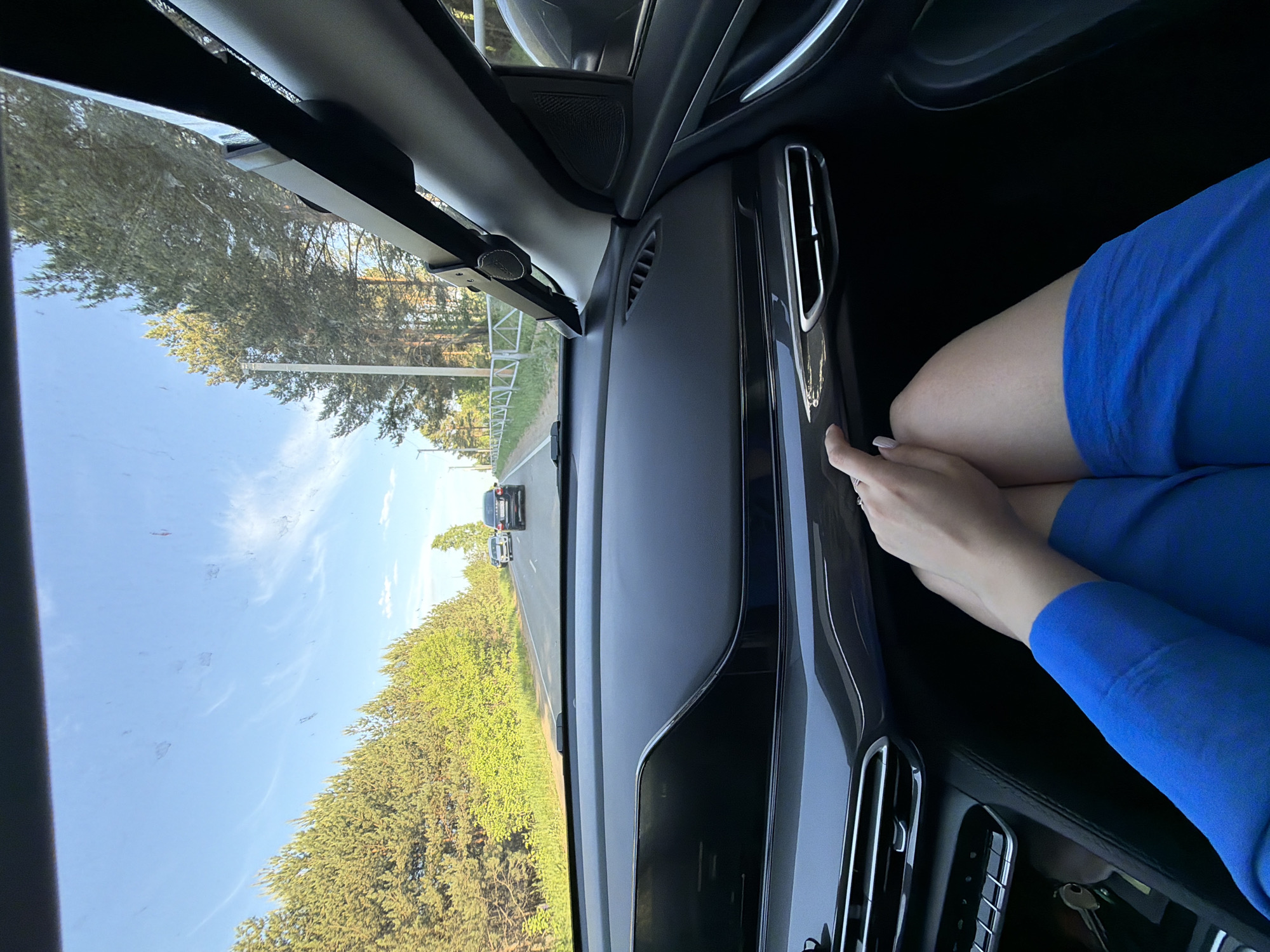 The image, taken from inside a car, shows a view of a road flanked by trees under a blue sky, suggesting a drive during daylight. In the foreground, part of a person wearing a blue garment rests their arm near the car's dashboard, which features air vents and other controls. The location appears to be outside Chaikovsky, Russia. Several cars can be seen on the road ahead. The image captures a moment during the car's journey.