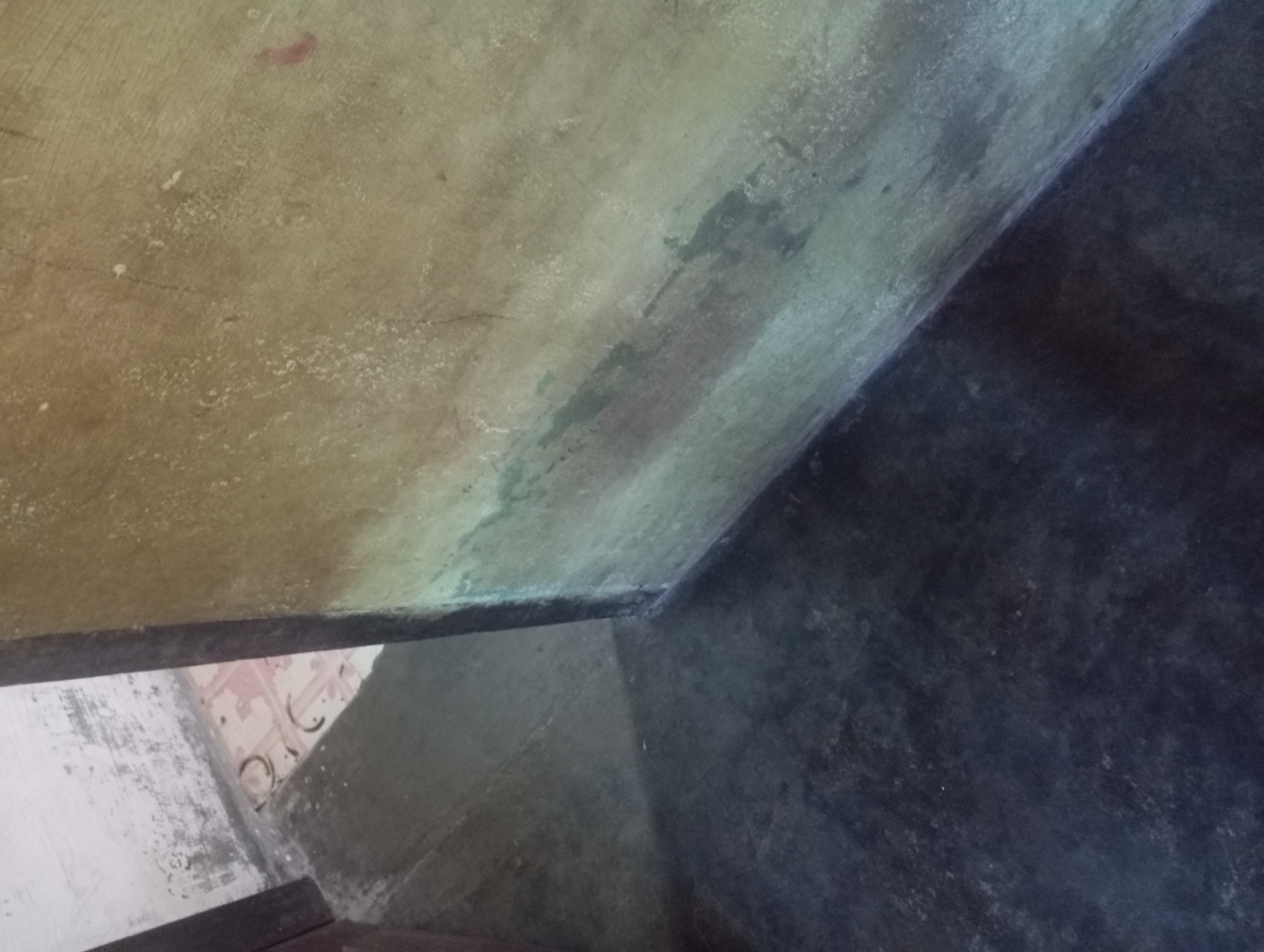 The image depicts a corner of a room, likely in Rejosari, Indonesia. The floor is a dark, weathered concrete, with a lighter grey concrete ramp leading towards a doorway. The wall is a textured, yellowish-brown material, showing signs of moisture damage with streaks of green and grey. To the left of the wall, a white wall with peeling paint is visible, and the floor underneath appears to be tiled with a pink and white pattern. The lighting suggests an indoor setting, possibly an unlit area or a room with dim natural light. There are no people or specific objects that would indicate a particular activity or event.