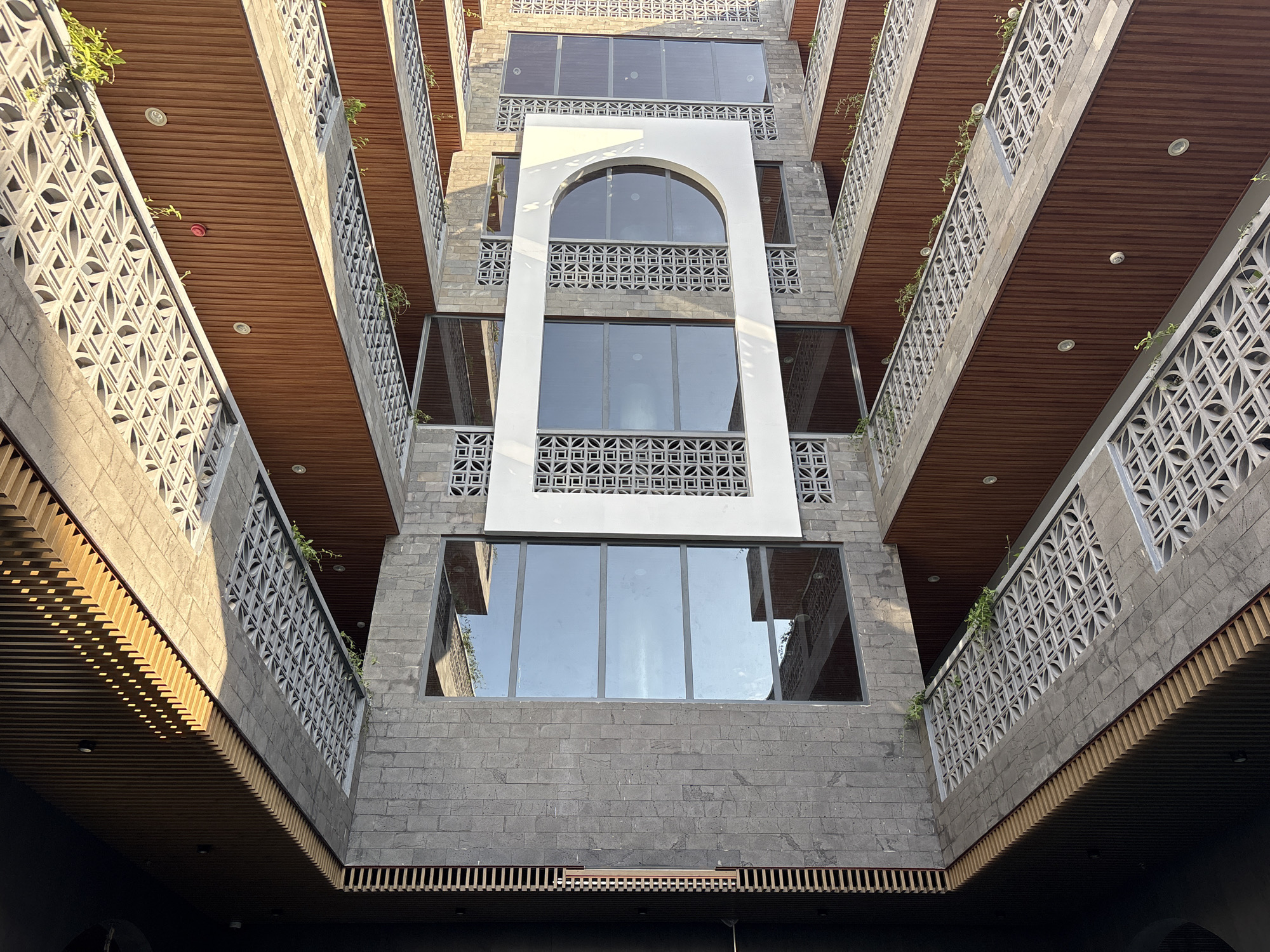 This image captures a striking architectural interior or courtyard of a multi-story building, likely in Đà Nẵng, Vietnam, viewed from a low angle looking upwards. The structure showcases a modern design blending robust materials with intricate patterns.
The primary building material for the walls is textured gray stone blocks, giving a solid, earthy feel. Contrasting with this are warm brown wooden slatted ceilings and the undersides of the balconies, which line both sides of the central void, creating horizontal lines and adding warmth. Each floor features balconies with decorative concrete block railings, displaying complex geometric patterns that enhance airflow and light diffusion. Small green plants are visible, cascading from some of these balconies, introducing organic elements to the structured environment. Recessed lighting fixtures are integrated into the wooden ceilings.
A prominent central section of the building is defined by a tall, white architectural frame, culminating in a large, elegant arched window at its apex. This white frame encloses multiple large, reflective windows that mirror the clear blue sky and the surrounding building elements. Below the arched window, rectangular windows are set into the gray stone, also showing reflections.
Bright sunlight casts sharp, distinct shadows across various surfaces, indicating a clear, sunny daytime, likely morning or late afternoon. No people or specific activities are visible, allowing the viewer to focus on the building's architectural details and play of light and shadow. The overall aesthetic is one of modern tropical design, characterized by its use of natural materials, emphasis on light, and thoughtful ventilation through the patterned blocks. No visible text is present in the image.