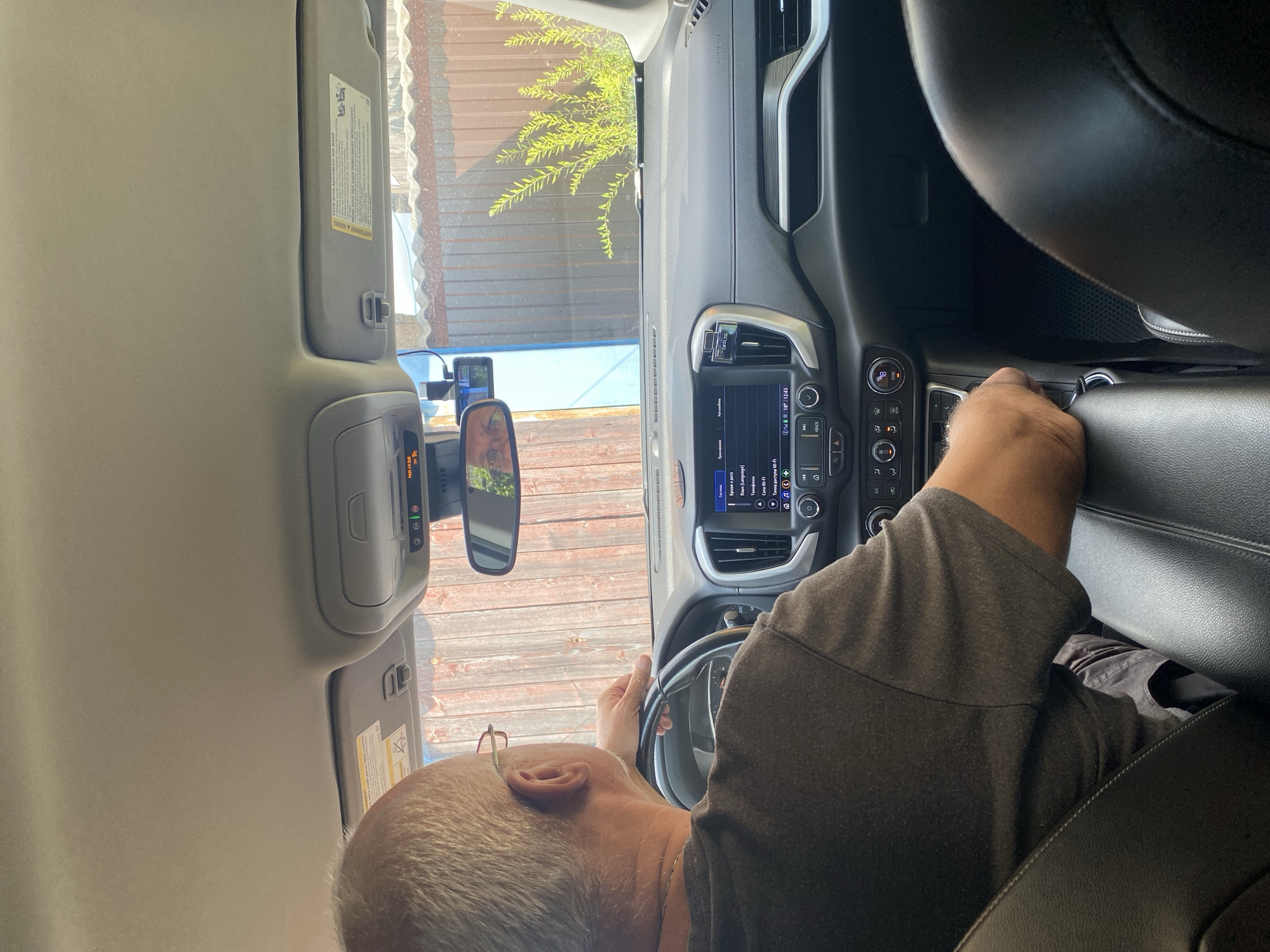 The image shows a man driving a car. He is wearing a dark t-shirt and glasses, and is holding the steering wheel. The car's dashboard is visible, with a touchscreen display showing information such as time, date, and Wi-Fi connectivity. The car appears to be on a road in Krupki, Belarus, as indicated by the outdoor environment with wooden fencing and greenery visible through the windows. The lighting suggests it is daytime.