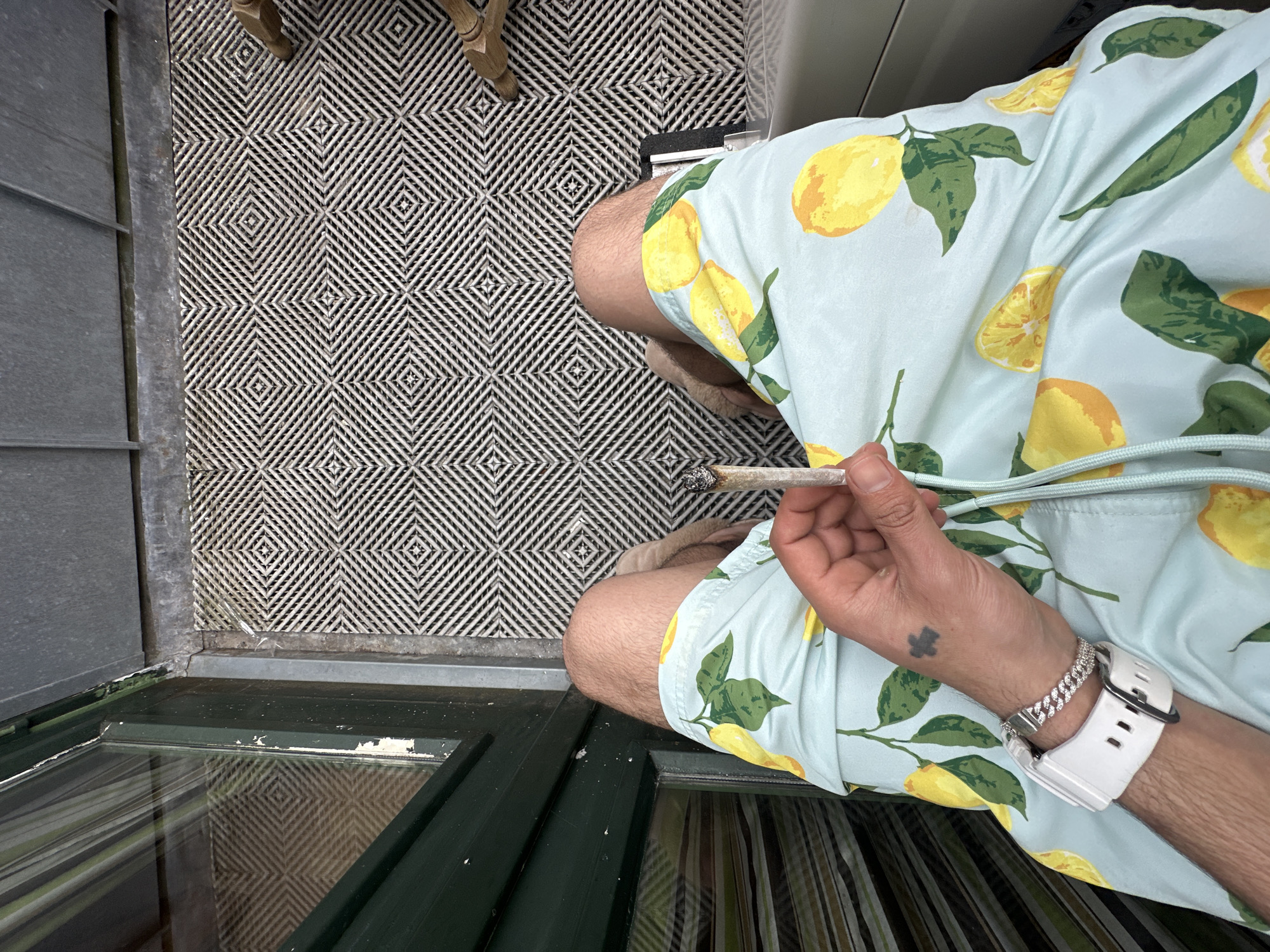 This image, taken from a high-angle perspective, captures a person seated or crouching in what appears to be a doorway or indoor threshold, likely in Amersfoort, Netherlands. The subject's legs, torso, and right arm are visible. They are wearing light blue shorts patterned with vibrant yellow lemons and green leaves.
On the person's right wrist, a white digital watch with a black buckle is worn, along with a shiny silver Cuban link-style bracelet. A small, dark tattoo resembling a four-leaf clover or abstract shape is visible on the back of their right hand. The person is holding a lit, hand-rolled cigarette, commonly referred to as a joint, between their thumb and index finger. The tip of the joint shows visible ash and a small glowing ember, indicating it is actively being smoked.
The floor is covered by a distinctive black and white geometric patterned mat or rug, composed of interlocking diagonal lines that form a series of squares. To the left of the person, a dark green door or frame with a glass panel is visible, reflecting a distorted view of vertical stripes, possibly curtains, from beyond the glass. A wooden furniture leg is partially visible in the upper background. The overall scene suggests a moment of casual relaxation. No visible text is present in the image.