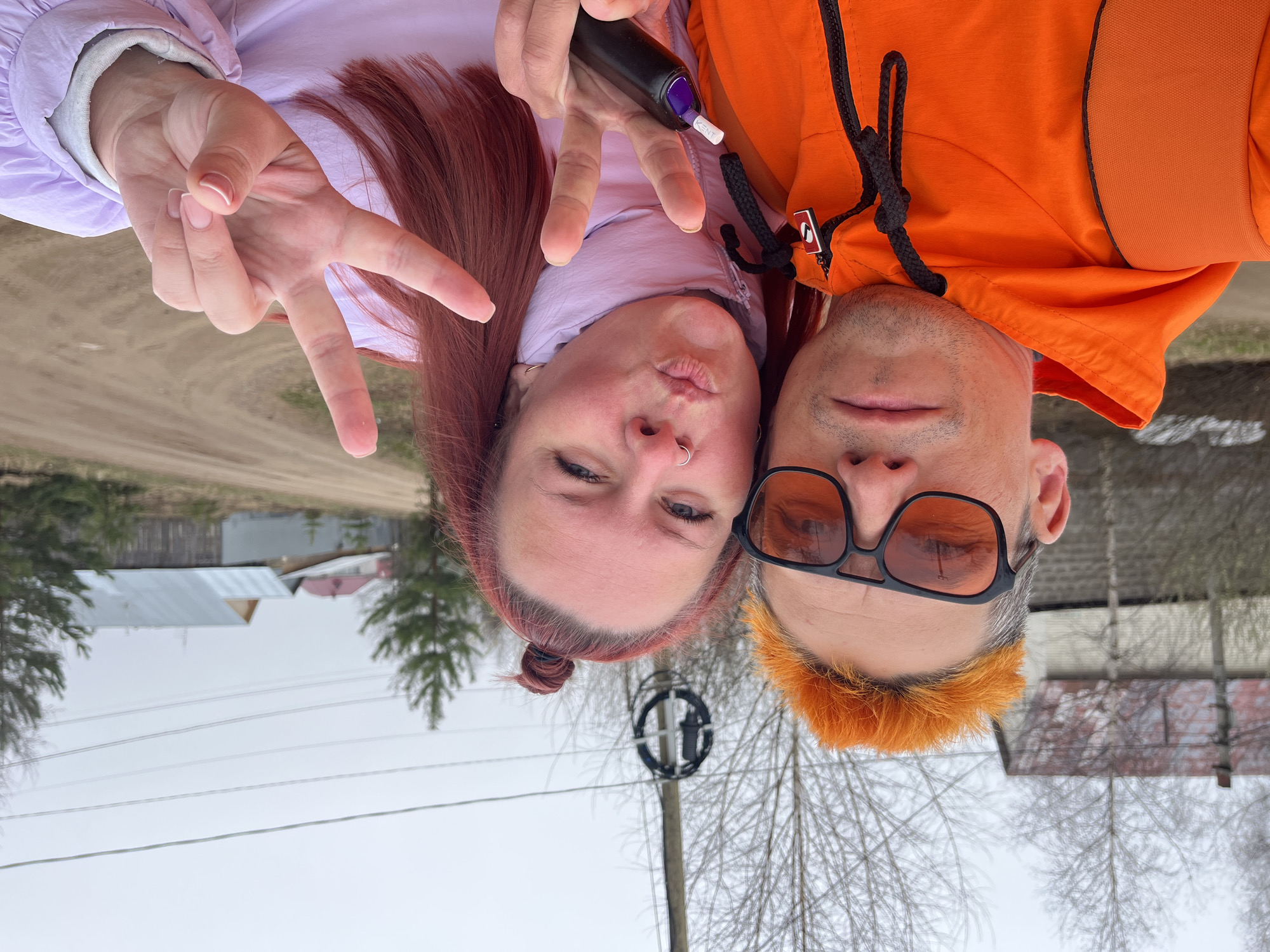Two individuals are posing for a selfie outdoors. A woman with auburn hair pulled back in a bun, wearing a lilac jacket, is making a peace sign with one hand and holding a black object with a "KENT" label in the other. Her lips are pursed as if giving a kiss. Beside her is a man with short, orange hair, wearing orange-tinted glasses and an orange hooded sweatshirt with black drawstrings.
The background is blurry, revealing a cold, overcast landscape with houses and bare trees. A utility pole with circular equipment is visible between the two people. In the distance, one can discern a dirt road and more buildings. The location is speculated to be in Chaikovsky, Russia, based on contextual clues. The image appears to be rotated 180 degrees.