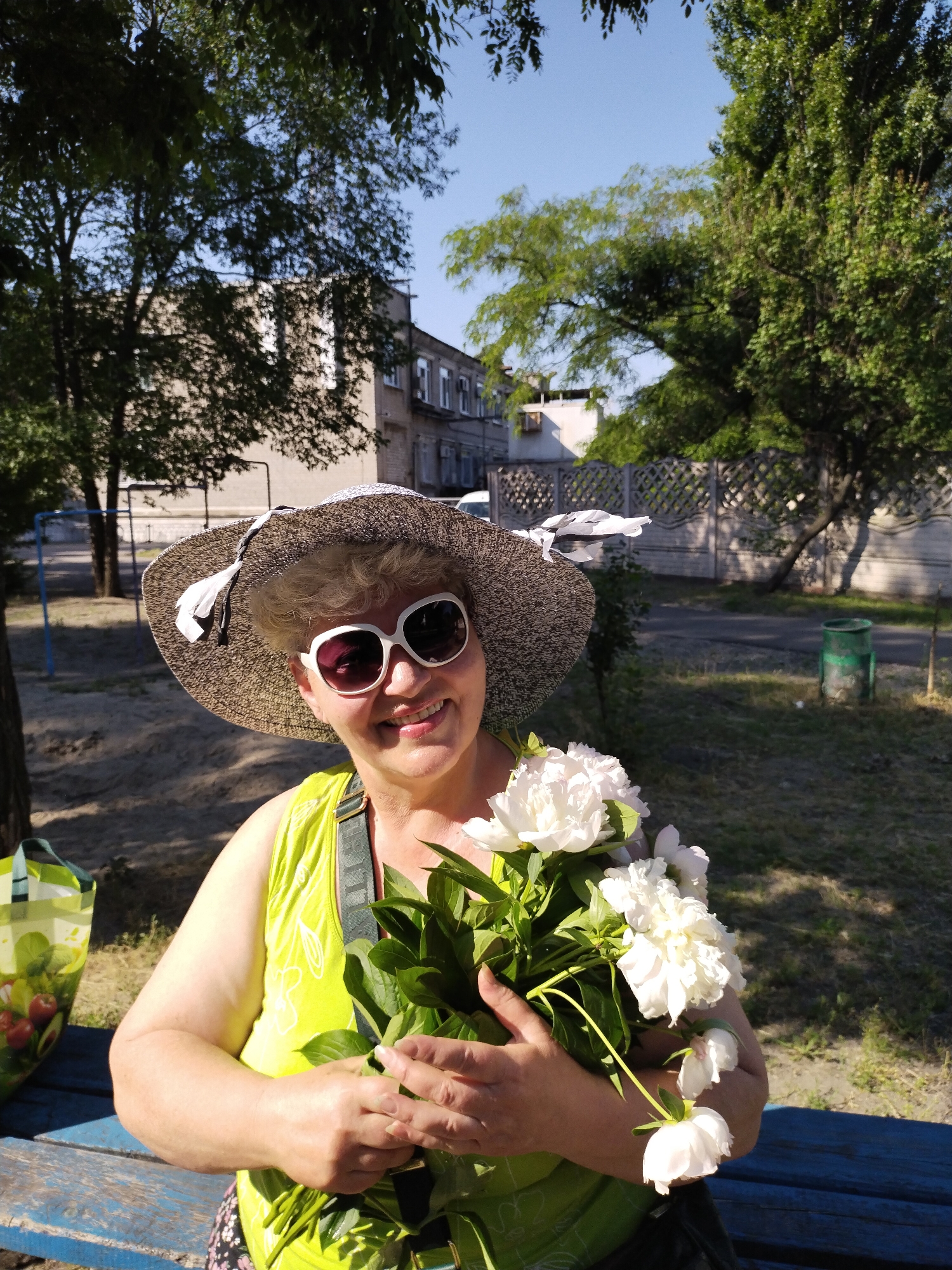 A woman in her 60s with blonde hair, wearing a wide-brimmed hat and large white sunglasses, smiles while holding a bouquet of white peonies. She is wearing a bright green sleeveless top and is seated on a blue wooden bench outdoors. In the background, a concrete fence, trees, and a multi-story building are visible. The scene appears to be a park or residential area in Horishni Plavni, Ukraine, judging by the presence of a children's swing set in the mid-ground and a shopping bag with produce imagery on the left. The lighting suggests it is a sunny day.