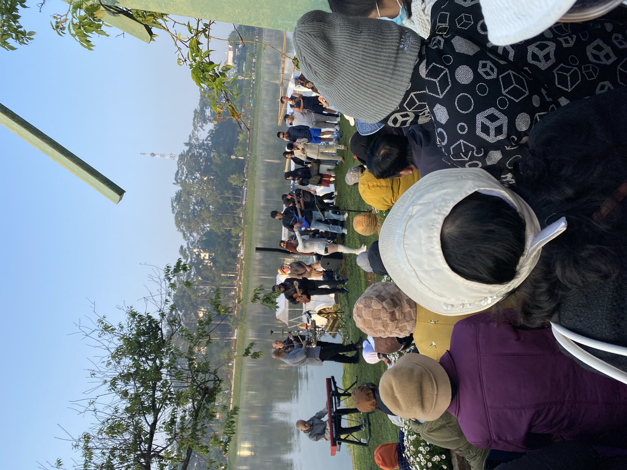 Here is a concise description of the image:
The image shows a lakeside setting, possibly in Da Lat, Vietnam, given the context clues. A group of musicians is performing along the waterfront. A crowd of people is gathered, likely watching the performance. The people are of varying ages and are dressed in casual attire, many wearing hats. A body of water is visible along the waterfront with a line of trees and buildings in the background. There is a tower structure in the distance, possibly a radio or communications tower. The perspective suggests the image was taken from an elevated position. The time of day is likely morning or late afternoon, as suggested by the soft lighting.