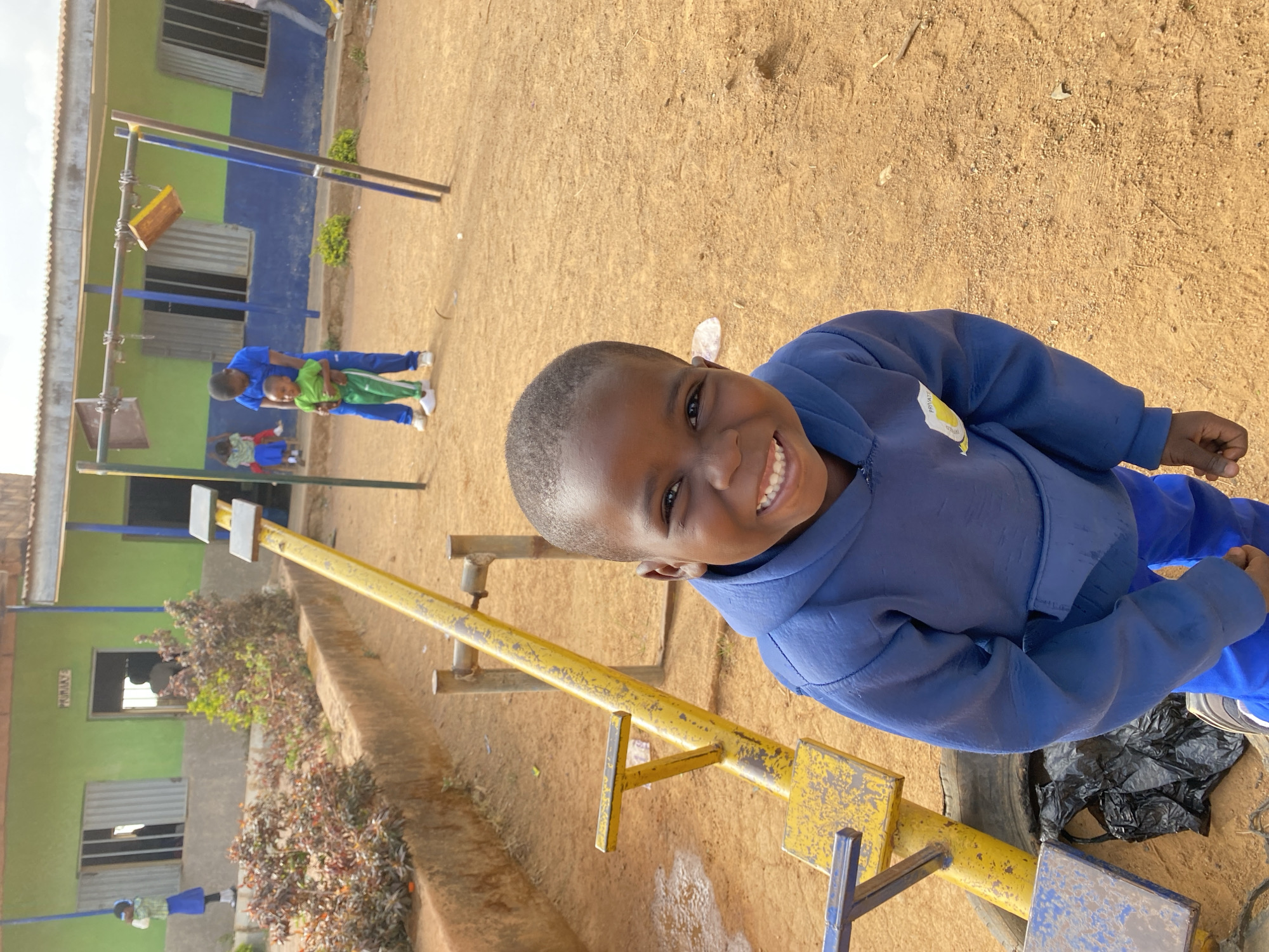 A young boy smiles broadly while standing next to a yellow seesaw in Gura Top, Nigeria. He wears a blue jacket and pants. In the background, other children and adults are visible near a green and blue building. The ground appears to be dirt or sand.