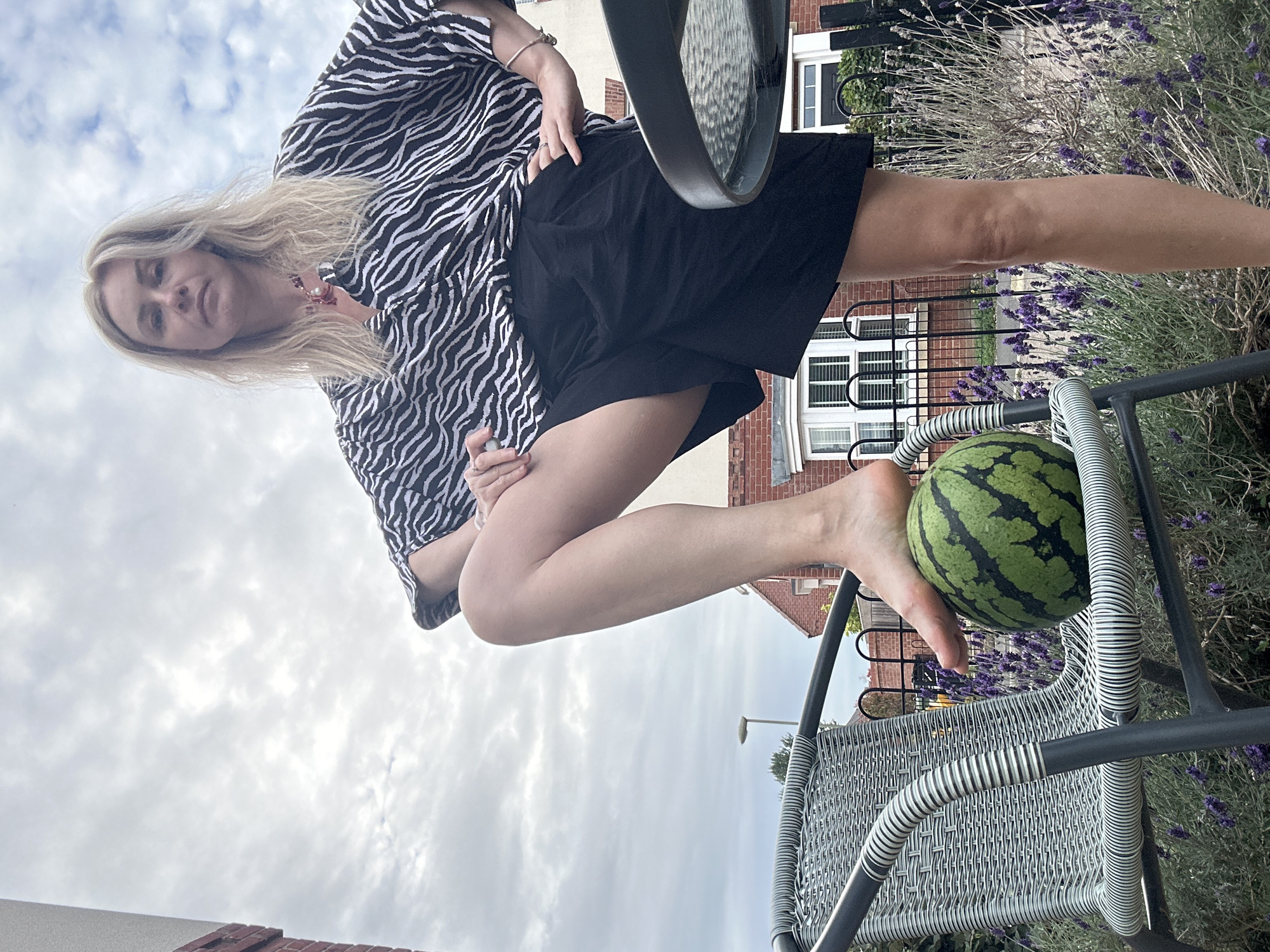 An outdoor, medium shot captures a woman with blonde hair, wearing a black and white zebra-print top and black shorts, standing with her right foot on a small watermelon resting on a wicker chair. The woman is positioned on the right side of the frame, with her gaze directed towards the camera. Her right leg is bent at the knee, with her foot pressing down on the watermelon. The chair, with a striped pattern on its arms, is on the left side of the image. In the background, a residential house with red brick and white window frames is visible, along with a garden featuring lavender plants and a black metal fence. The sky above is cloudy.