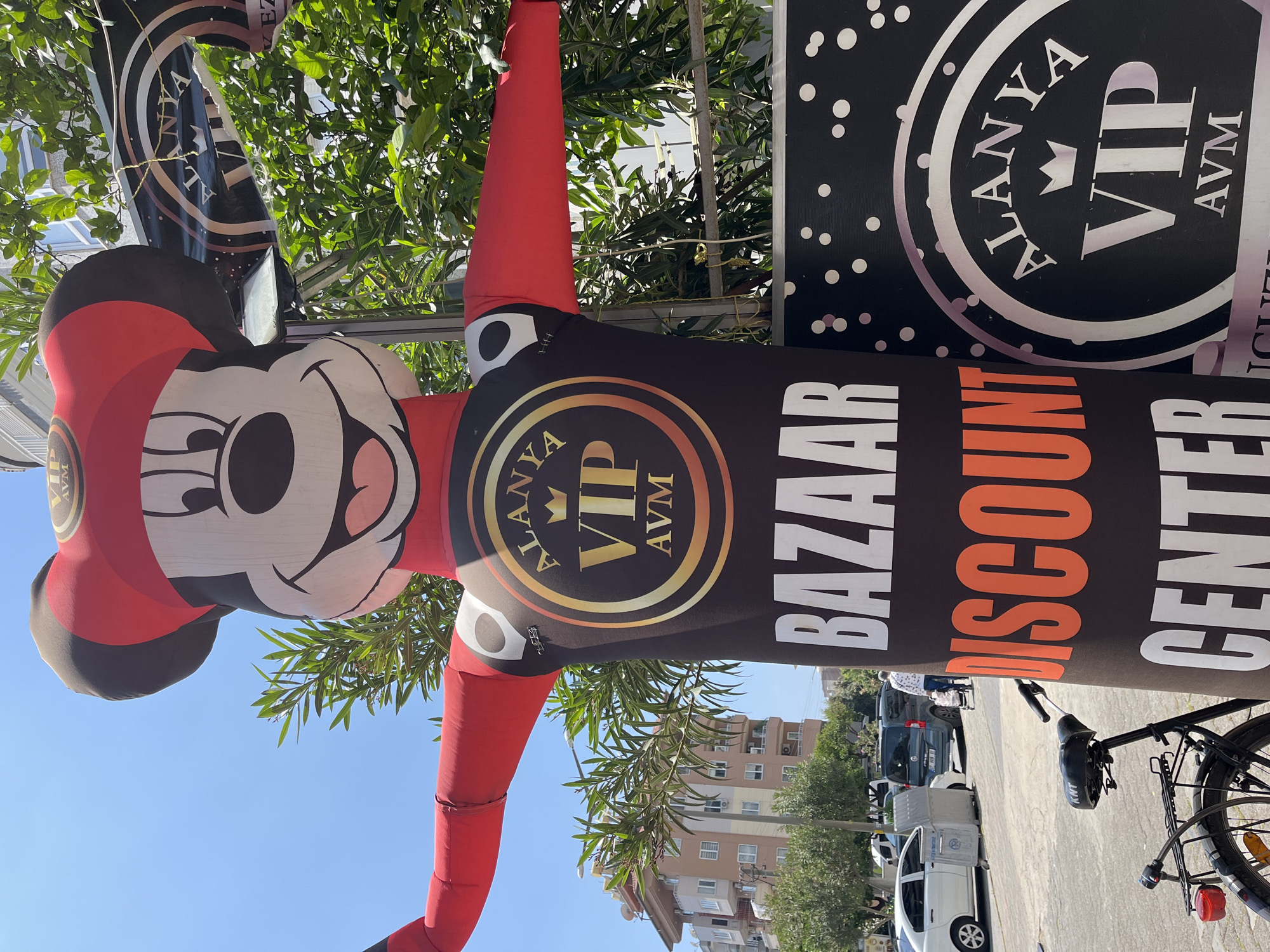 The image displays an outdoor street scene in Alanya, Turkey, bathed in bright daylight under a clear blue sky. The main subject is a large, vertical inflatable character, stylized to resemble Mickey Mouse, dressed in red and black. On its chest, a gold circular logo reads "ALANYA VIP AVM".
Adjacent to the inflatable stands a tall, black banner or sign. This sign also prominently features "ALANYA VIP AVM" within a large silver circle, set against a background of white polka dots. Below this logo, the words "BAZAAR" (in white), "DISCOUNT" (in orange), and "CENTER" (in white) are stacked vertically, advertising a commercial establishment.
In the background, abundant green foliage from trees suggests a pleasant, possibly Mediterranean, environment. Beyond the greenery, several multi-story buildings with numerous windows and balconies line what appears to be a street. A dark-colored car and a white utility vehicle are parked on the street. A black bicycle with a rear reflector is visible in the foreground, near the base of the sign. The scene presents a static view of a commercial area, likely in a town or city, with the inflatable and signs acting as prominent advertisements. No discernible human activity is taking place in the immediate foreground.