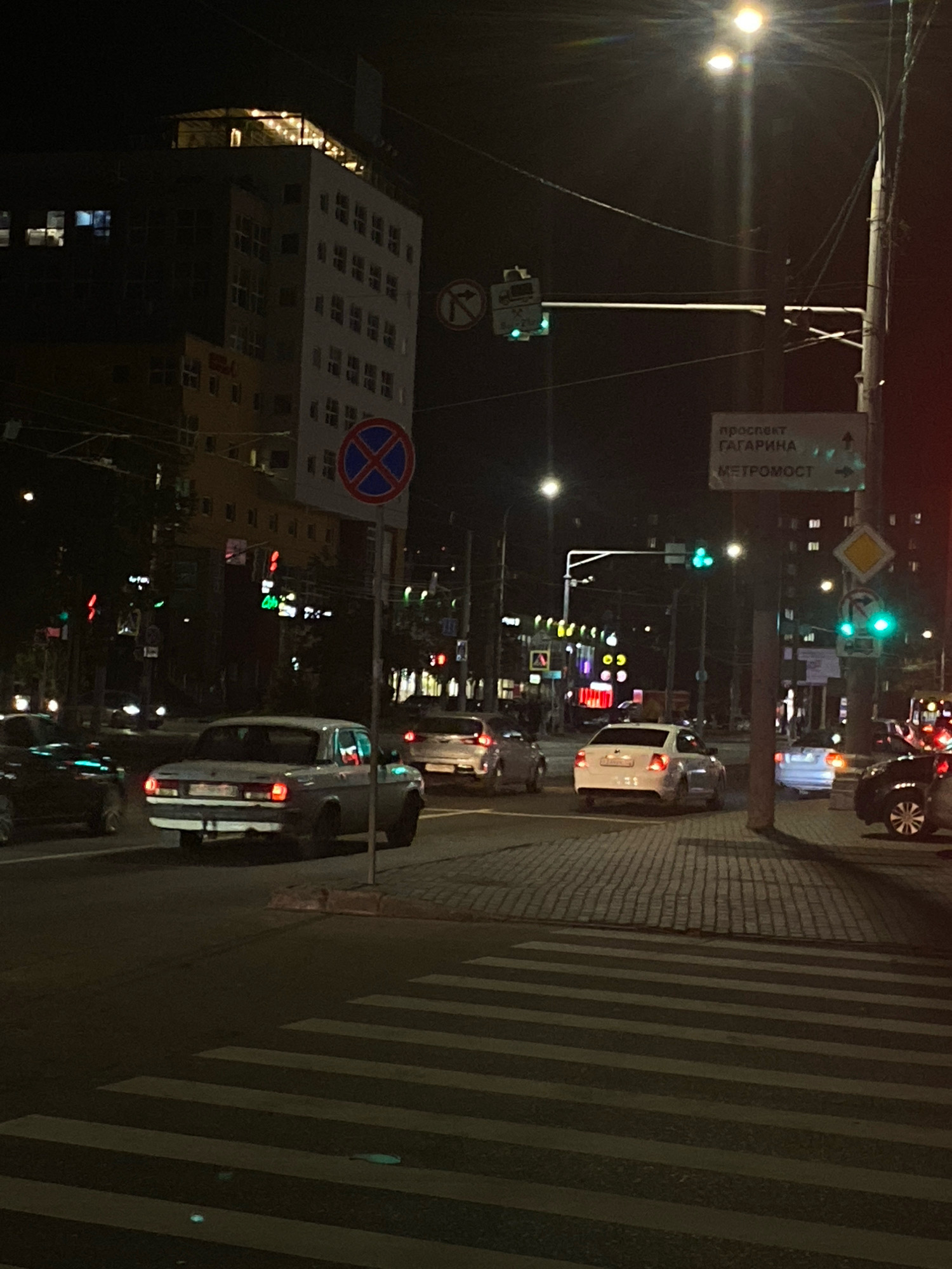 The image depicts a nighttime street scene in Nizhny Novgorod, Russia, likely on Gagarin Avenue near the Metromost bridge. Several cars are visible, including a light-colored sedan in the foreground facing left and a white sedan further down the street. Traffic lights are illuminated, with green lights showing. A pedestrian crossing with white stripes is prominent in the lower part of the image. Buildings line the street, with one prominent multi-story building featuring illuminated windows and a rooftop structure. Streetlights and power lines are also present. A road sign indicating directions to "Prospect Gagarin" and "Metromost" is mounted on a pole. The overall atmosphere is urban and busy, characteristic of a city at night.