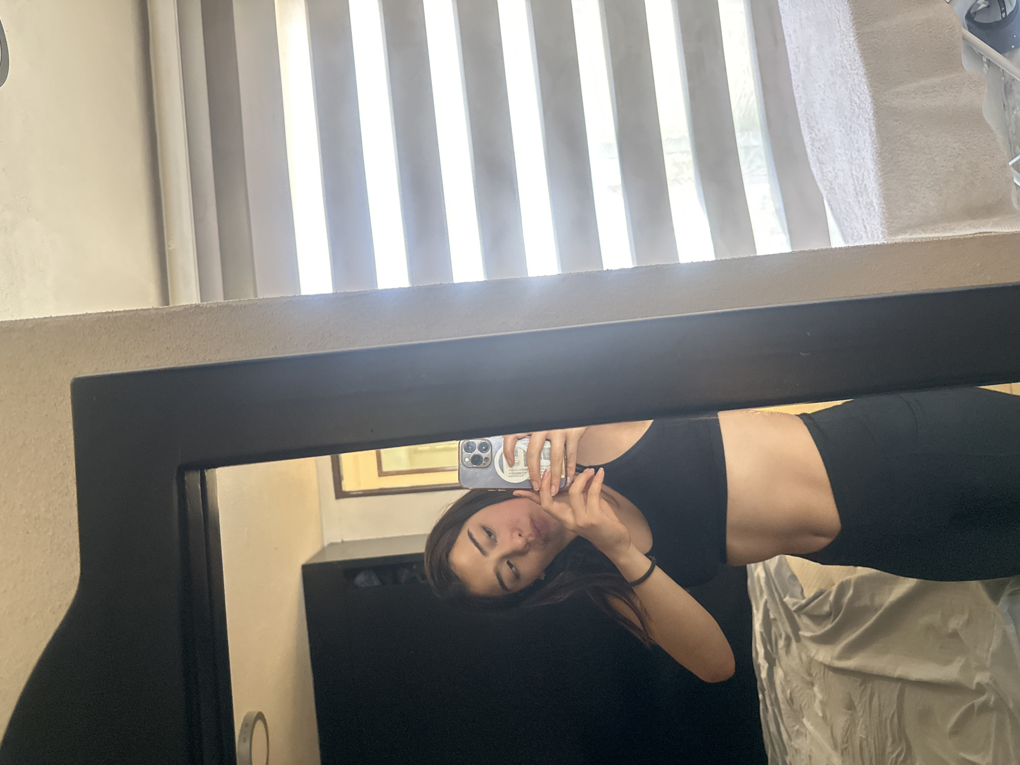 An image captures a young woman taking a mirror selfie in an indoor setting, likely a room in Puebla, Mexico. The woman, appearing in her late teens or early twenties, is reflected in a large mirror with a thick, dark frame. She is wearing a black sleeveless crop top and a matching black skirt or shorts. She holds a smartphone, possibly an iPhone with multiple rear cameras and a light-colored case, in her right hand to capture the image. Her dark hair falls around her shoulders, and she poses with her left hand near her face, looking directly into the camera/mirror.
Above the mirror, representing the real-world background, bright daylight streams through vertical blinds or slats, suggesting it is daytime. The wall above the mirror has a visible texture. On the right side of the real background, a white, textured object, possibly a towel or soft furnishing, is partially visible. Within the mirror's reflection, behind the woman, a dark piece of furniture (possibly a dresser or cabinet) and a light-colored wall with a framed item are discernible. No specific city landmarks or legible text are visible in the image itself, beyond the general location context provided. The woman's expression is neutral and direct.