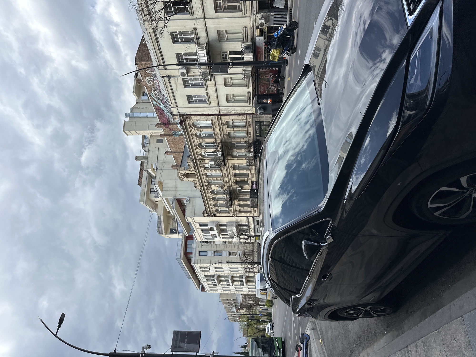 The image shows a street in Tbilisi, Georgia, lined with buildings of varying architectural styles, including ornate facades with balconies. A black SUV dominates the foreground, its glossy surface reflecting the sky and surrounding buildings. A streetlight and street sign are visible on the left side of the road. In the background, traffic includes cars and a scooter. The sky is overcast with clouds.