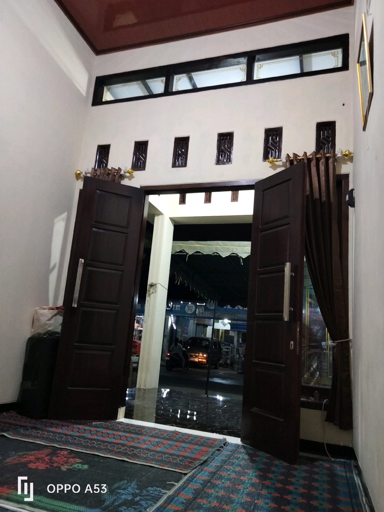 The image shows a hallway with two dark brown wooden doors, one slightly ajar. The doors have silver handles. On the left, a large black speaker is visible, with a white and red bag placed on top of it. The floor is covered with patterned rugs, predominantly in dark colors with red and blue accents. In the background, through the open doorway, a street scene is visible at night, with a dark-colored van parked and a person on a motorcycle. There are also shops with awnings. The walls are painted a light color, and the ceiling is a dark reddish-brown. Small, decorative windows are set high on the wall. The image is dated "OPPO A53". The scene is likely a residence or establishment in Trenggalek, Indonesia, given the text overlay.