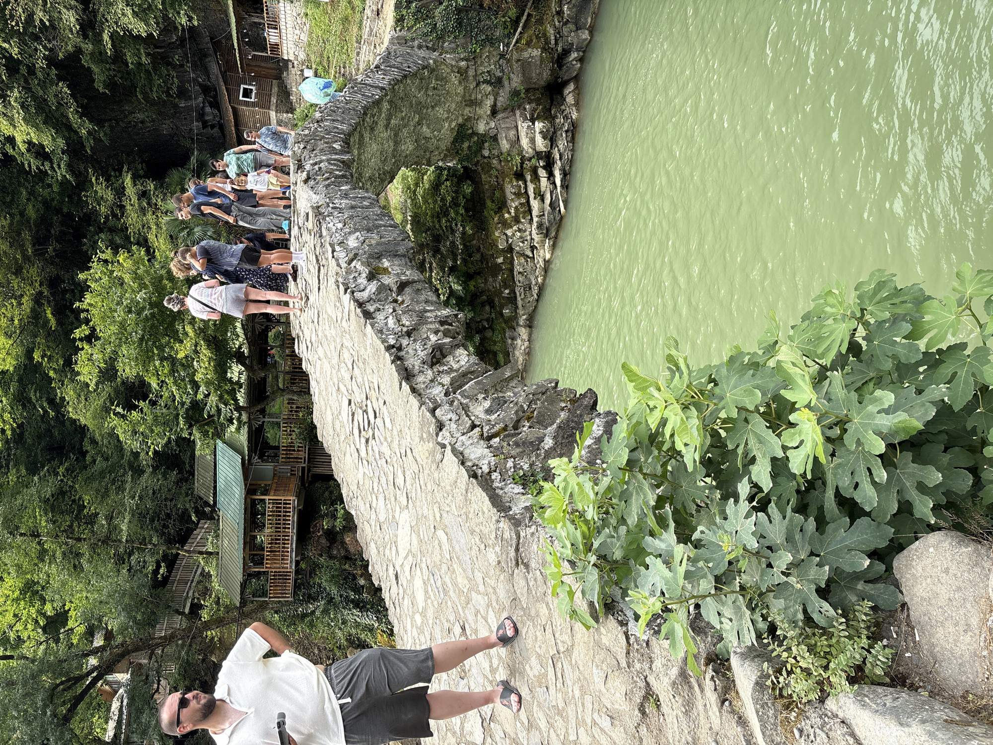 A man stands on a stone bridge in a lush, green environment. He is wearing sunglasses, a white polo shirt, and gray shorts. He holds a phone in his left hand and has his right hand on his hip. A group of people are walking across the bridge in the background. The bridge spans over a body of greenish water. There are buildings nestled in the trees on either side of the river. The foliage is dense and green.