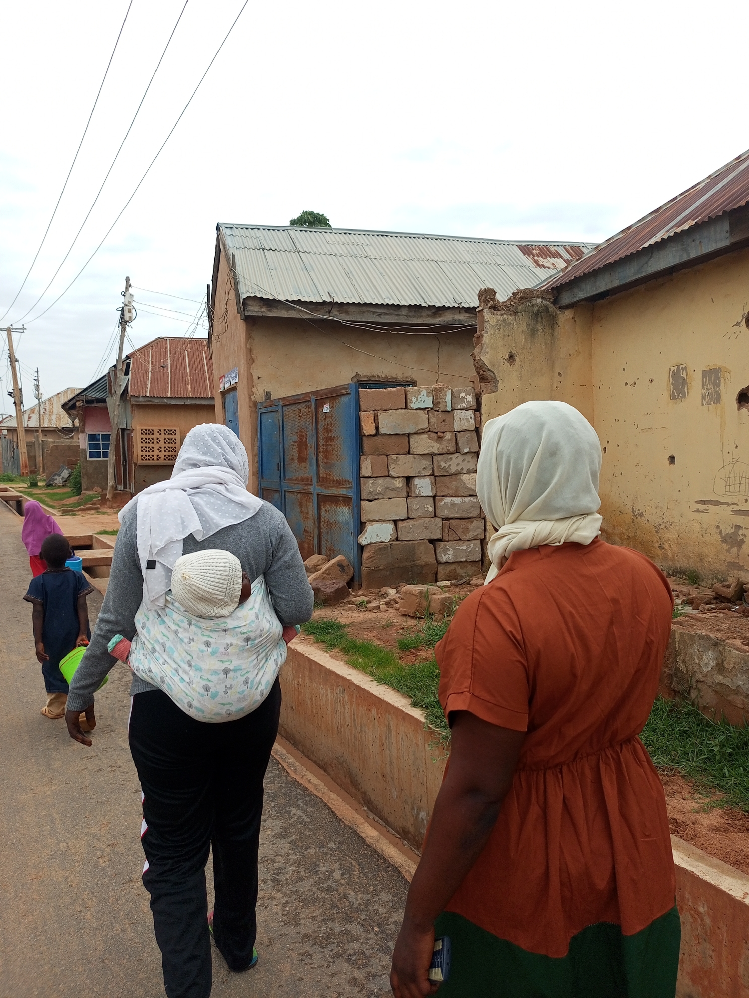 In Gombe, Nigeria, on a paved road lined with a low concrete barrier and sparse vegetation, three individuals are walking away from the camera. The foremost person, a woman, wears an orange dress and a light-colored headscarf. She is followed by another woman with a grey sweater and a white headscarf, carrying a baby on her back in a patterned wrap. The baby is wearing a white knitted cap. A young child, wearing a dark blue shirt and carrying a green bucket, walks ahead of the two women. In the background, a row of buildings with corrugated metal roofs and light-colored, aged walls are visible, some showing signs of disrepair. One building features a blue metal door and stacked bricks. Overhead, electrical wires stretch across a cloudy sky.