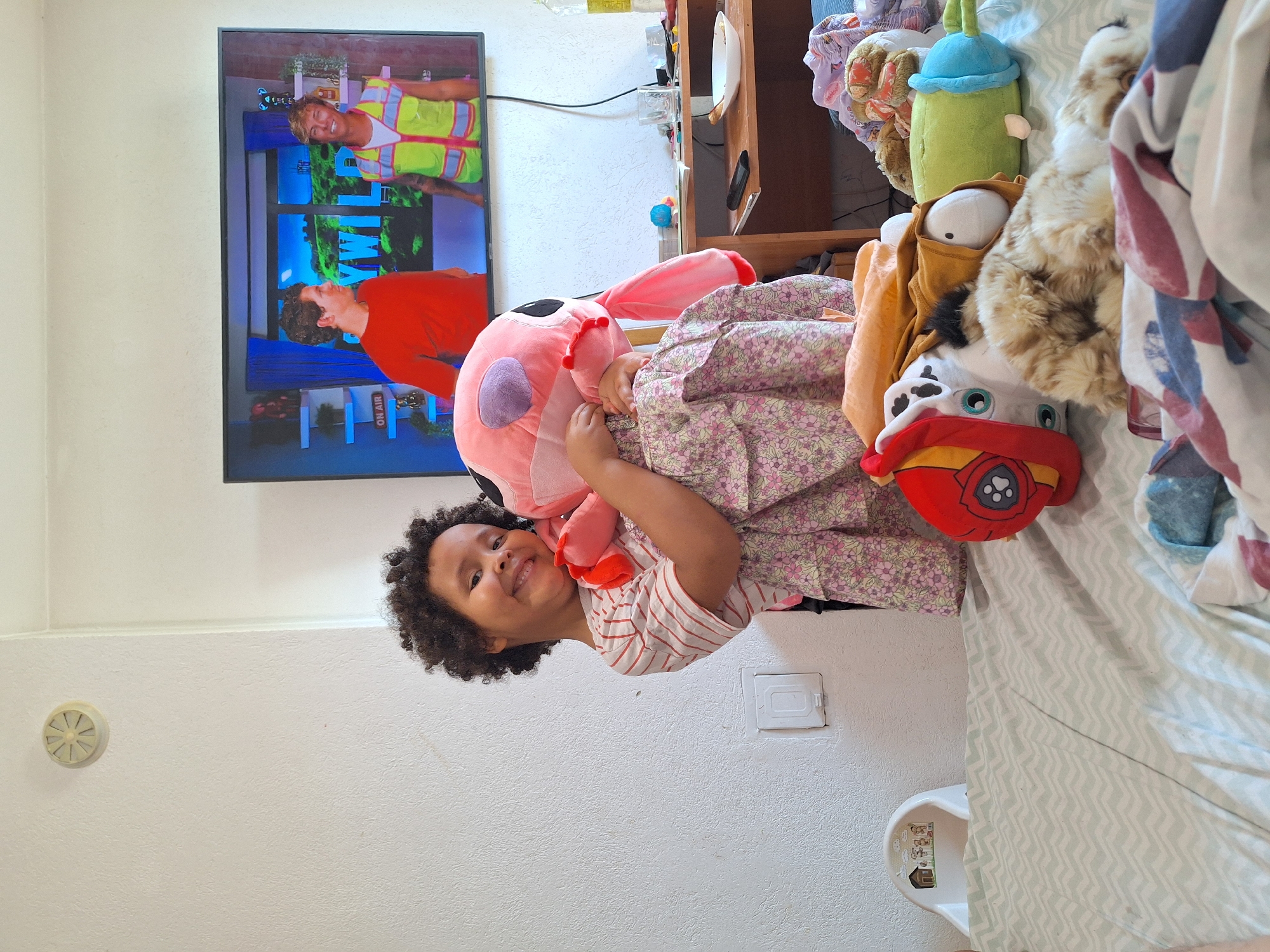 A young girl with curly dark hair is holding a large pink stuffed animal and smiling at the camera. She is wearing a striped t-shirt and a floral print dress. In the background, a television screen shows two men, one in a red shirt and the other in a yellow vest. A table with various items is visible to the right of the television. The scene appears to be indoors, likely a bedroom or living room, with a bed in the foreground covered in blankets and stuffed animals. The location is Blagoevgrad, Bulgaria.