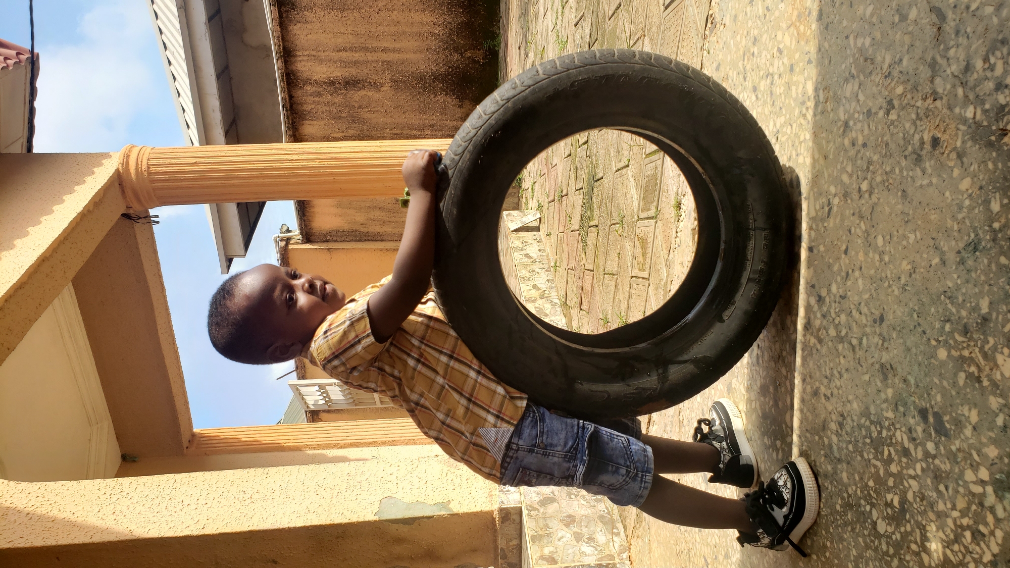 A young child stands in front of a large tire outdoors in Sabon Tasha, Nigeria. The child is wearing a yellow and brown plaid short-sleeved shirt and blue denim shorts, along with black and white sneakers. The child is leaning on the tire with their right hand. The tire is black and appears to be weathered. The background consists of a building with yellow-painted walls and columns, and a tiled floor. The sky is visible above the building. The overall scene suggests a residential area in Sabon Tasha, Nigeria.